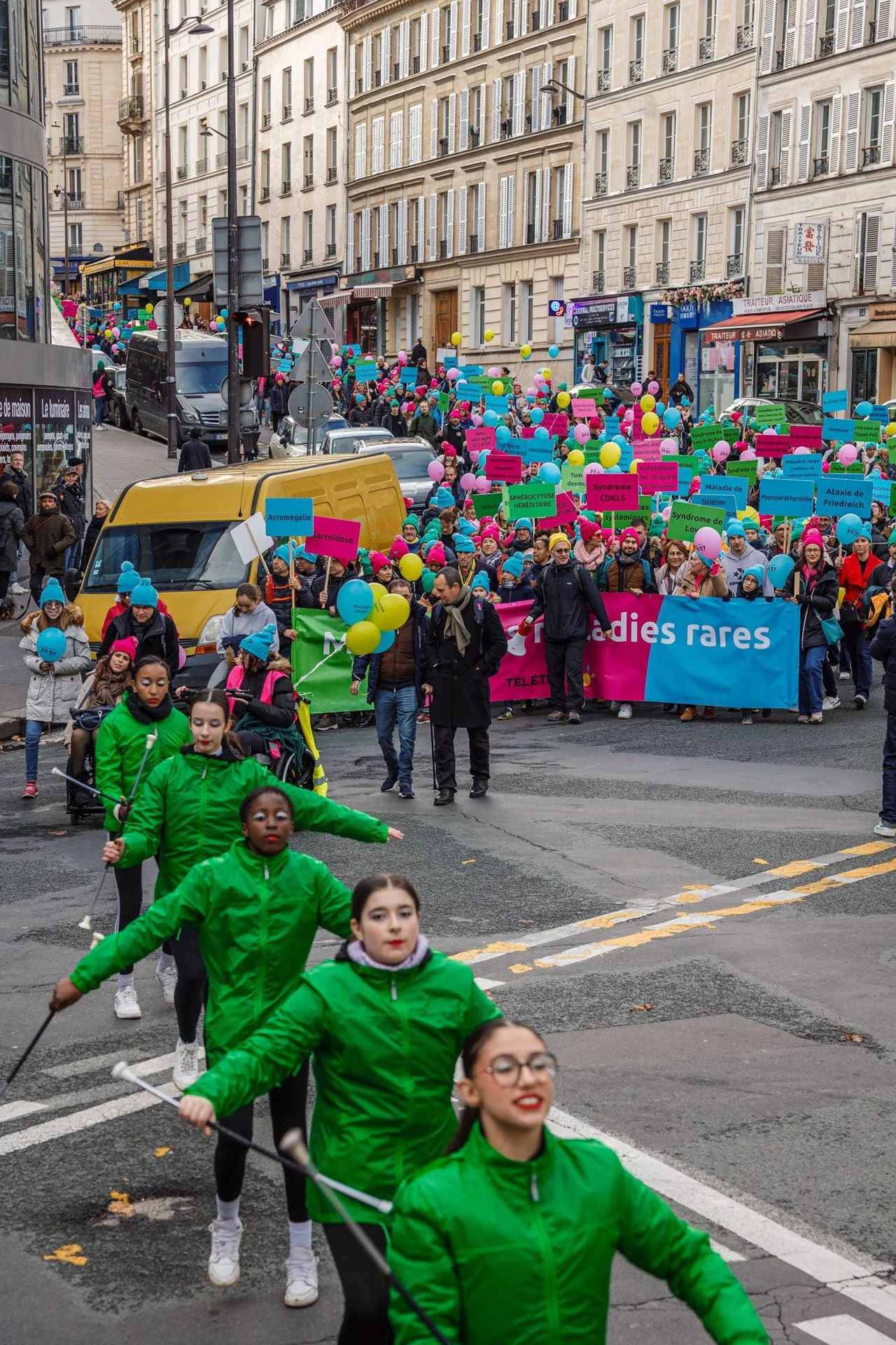 Manifestation dans une rue urbaine avec des participants portant des chapeaux roses ou bleus, tenant des ballons colorés et des pancartes sur le thème des maladies rares, avec des bâtiments résidentiels en arrière-plan.