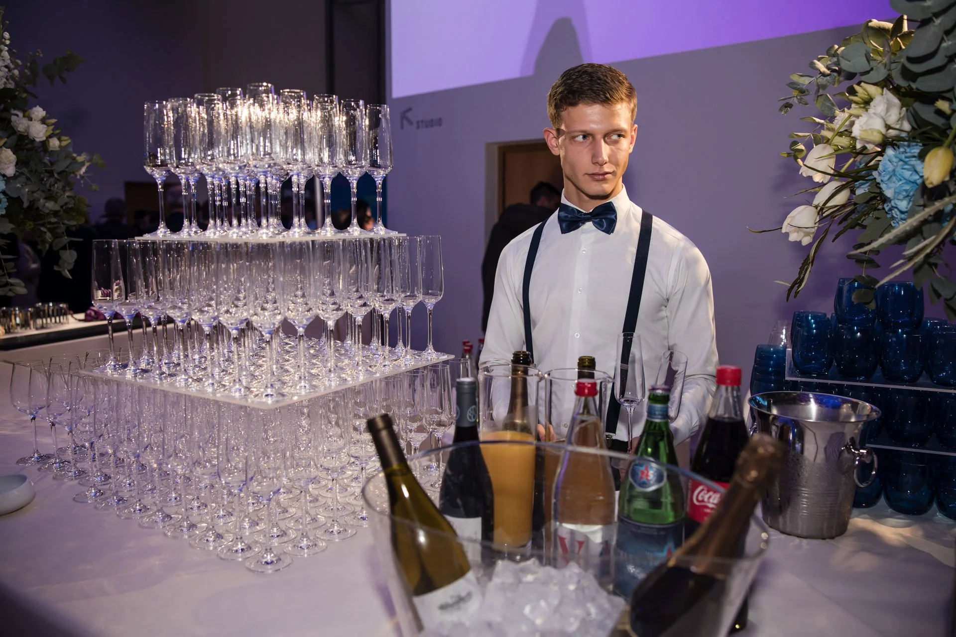 Un jeune homme en costume de serveur avec un nœud papillon se tient derrière un bar à boissons avec une pyramide de verres à champagne vides, diverses bouteilles et éléments pour préparer des cocktails, dans une ambiance de réception ou de fête.