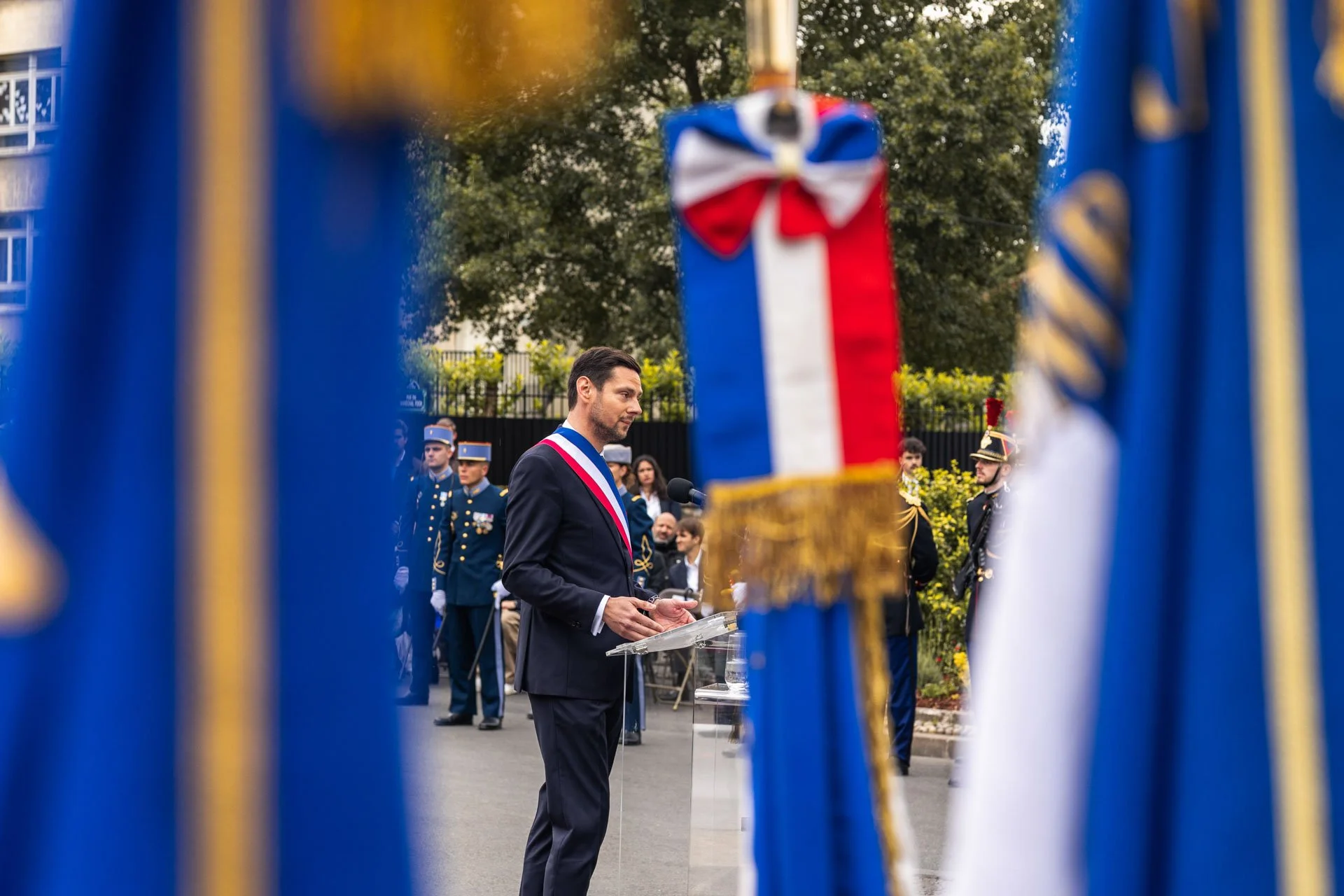 Un homme en costume noir avec une banderole tricolore parle lors d'une cérémonie, entouré de militaires habillés en uniforme, vus à travers une clôture décorée de rubans bleus et jaunes.