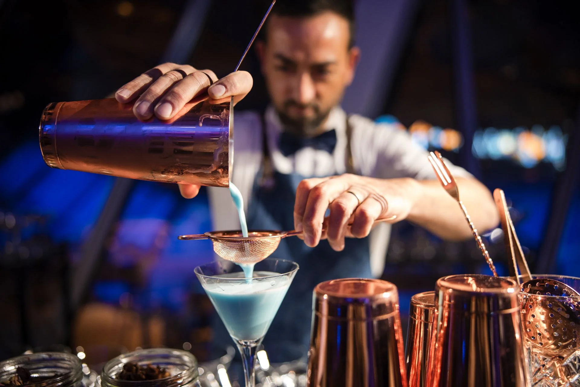 Barman prépare une boisson dans un verre à cocktail, entouré d'ustensiles en cuivre, avec un fond flou de lumières colorées.