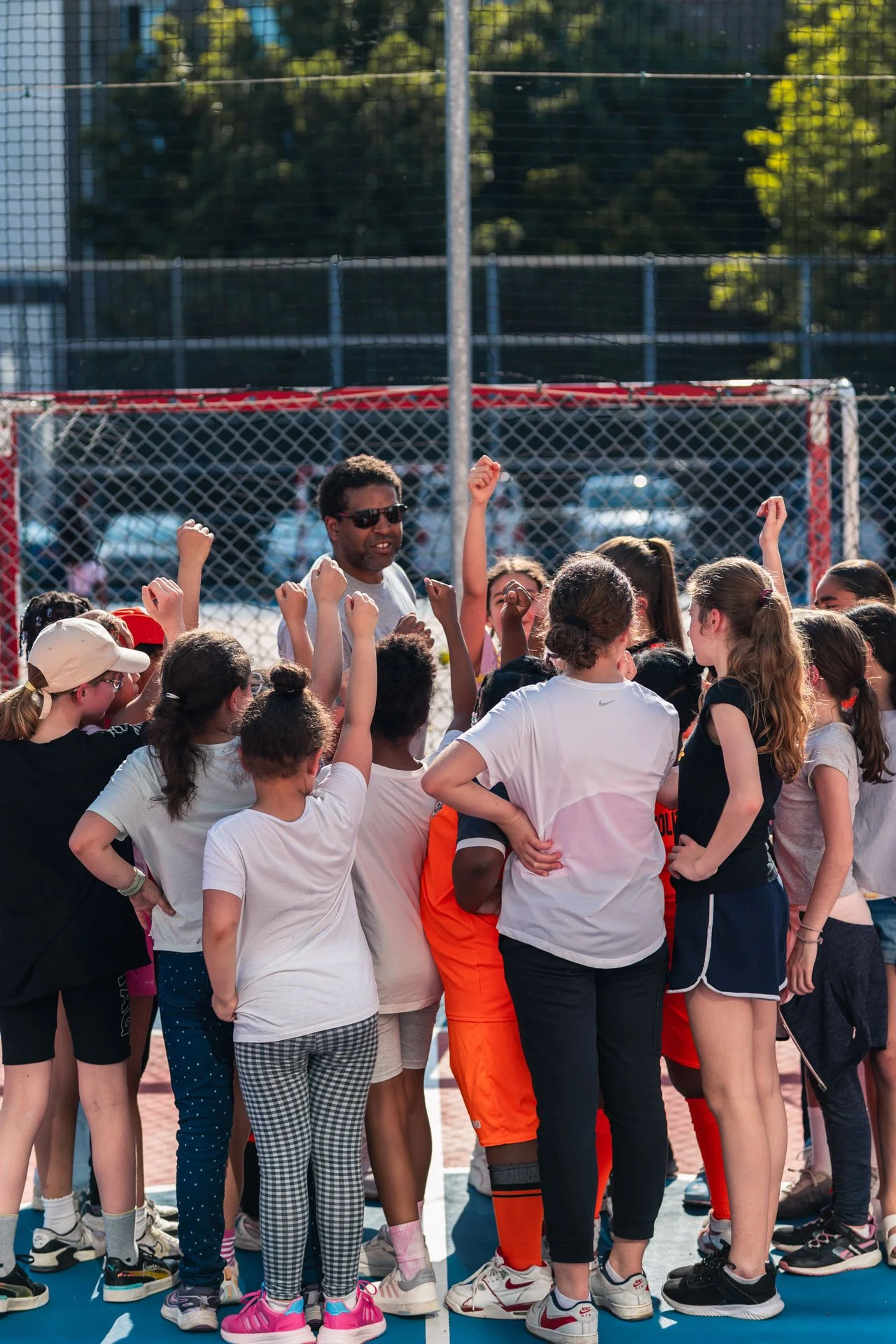 Un groupe d'enfants et une jeune adulte en train de se rassembler autour d'une personne adulte, probablement un entraîneur ou un coordinateur, sur un terrain de sport en plein air, ensoleillé avec un fond d'arbres et de clôtures de sport.