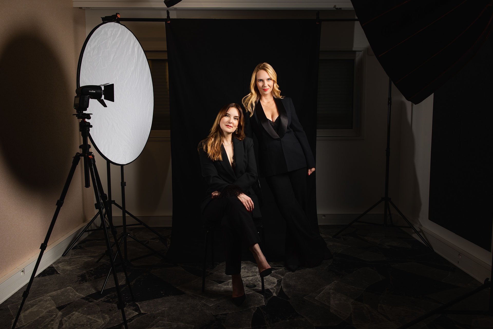 Deux femmes en tenue noire posent dans un studio de photographie avec un fond noir, un éclair de studio et un réflecteur blanc.
