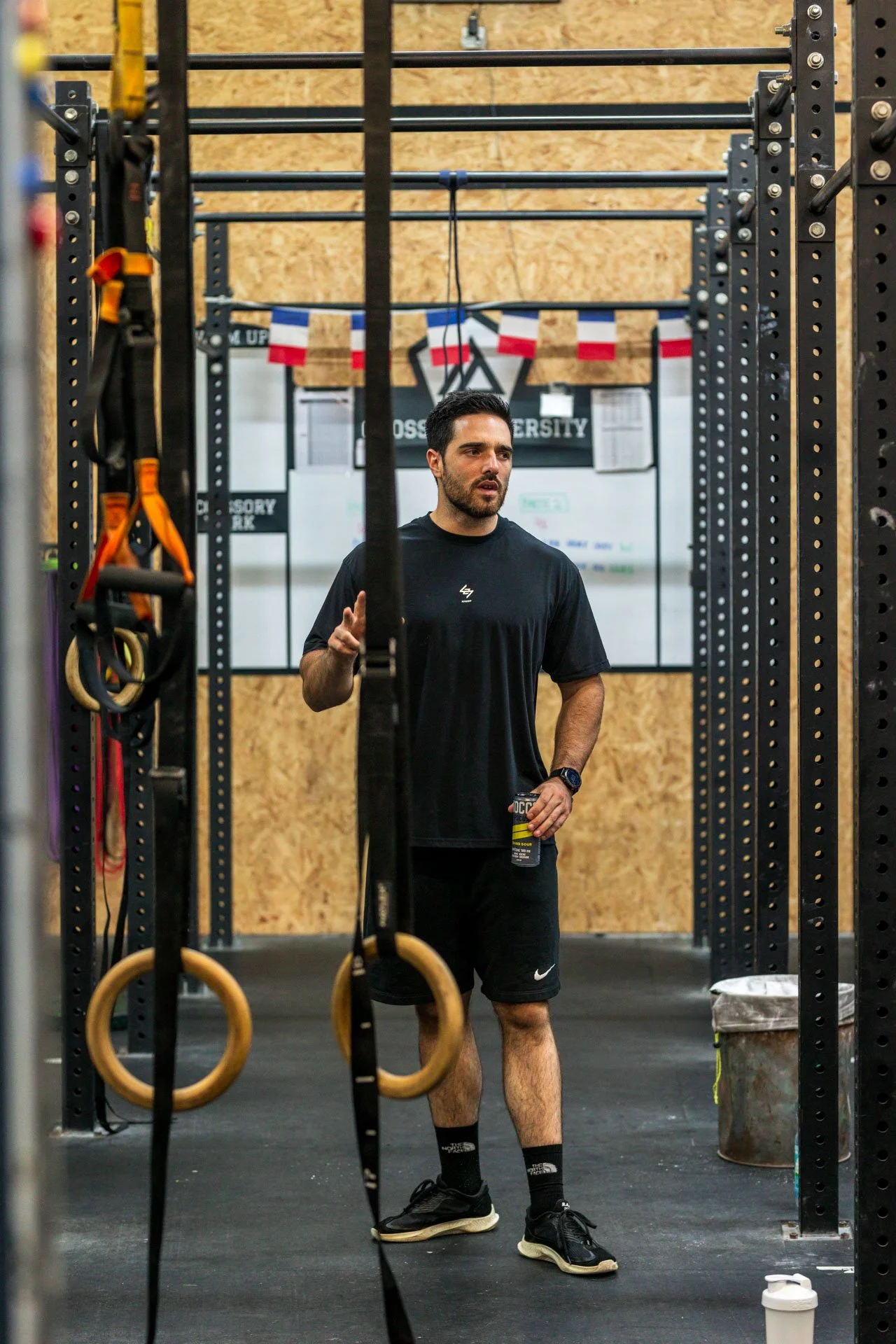 Un homme dans une salle de sport, portant un t-shirt noir et des shorts noirs, tient une boisson en main. Il semble prêt à faire de l'exercice, entouré d'équipements de musculation et de pognée de gymnastique.