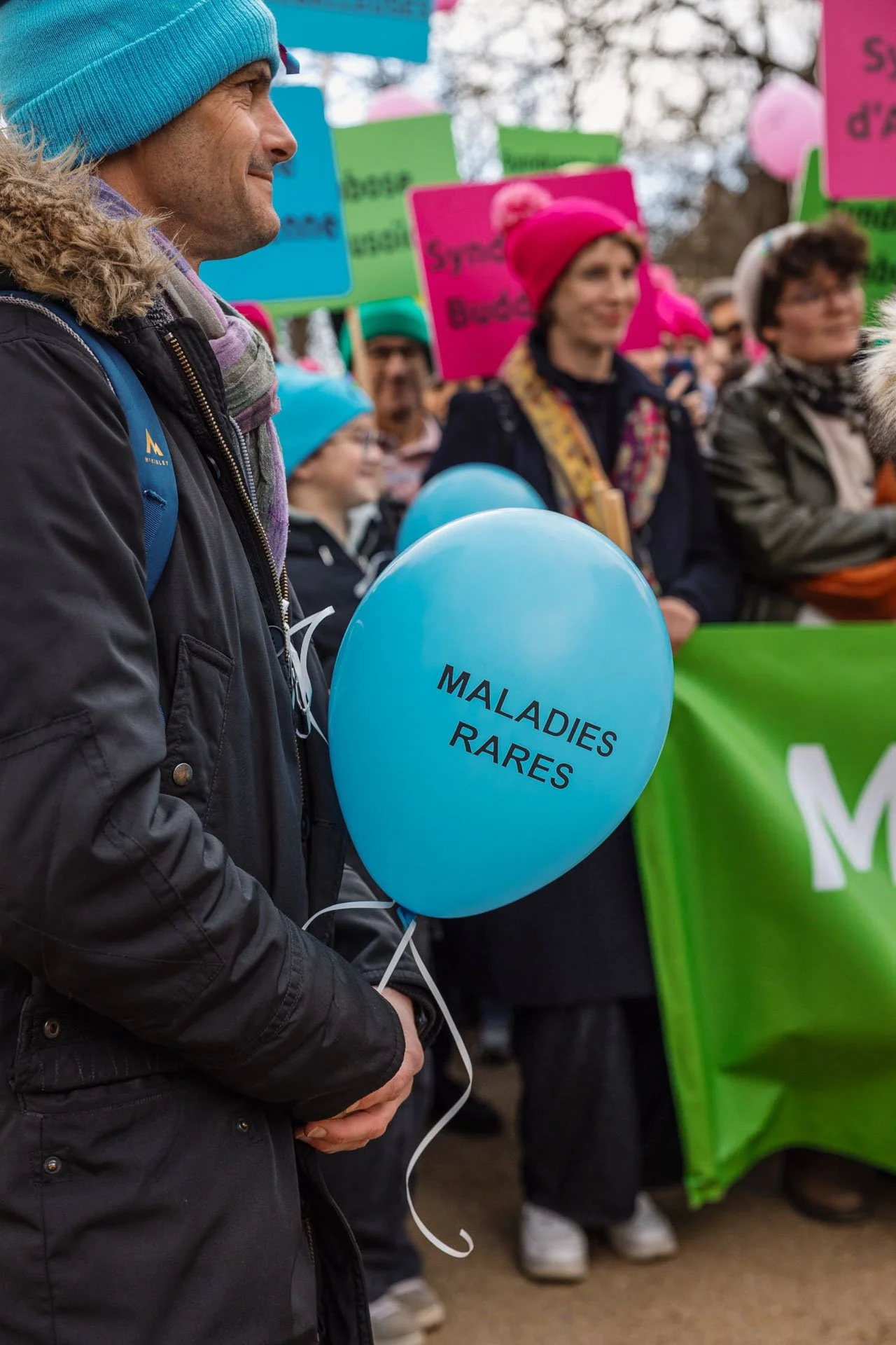Une personne tenant un ballon bleu avec l'inscription "MALADIES RARES" lors d'une manifestation ou un rassemblement, avec d'autres personnes en arrière-plan portant des pancartes colorées.