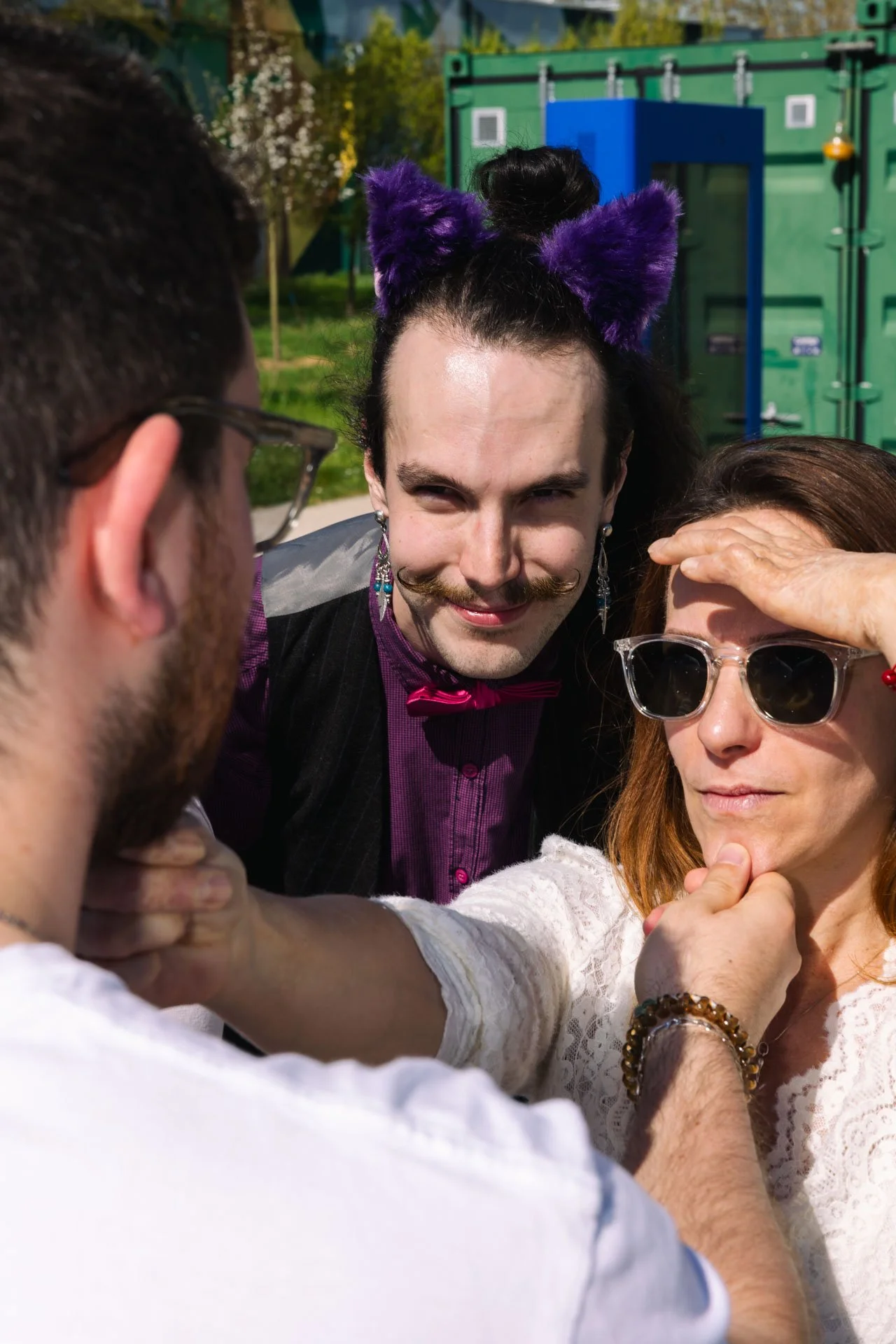 Groupe de trois personnes. Une femme avec des lunettes et des bijoux, un homme avec des oreilles de chat violettes et un homme vu de dos avec des lunettes, tous en train de poser pour une photo