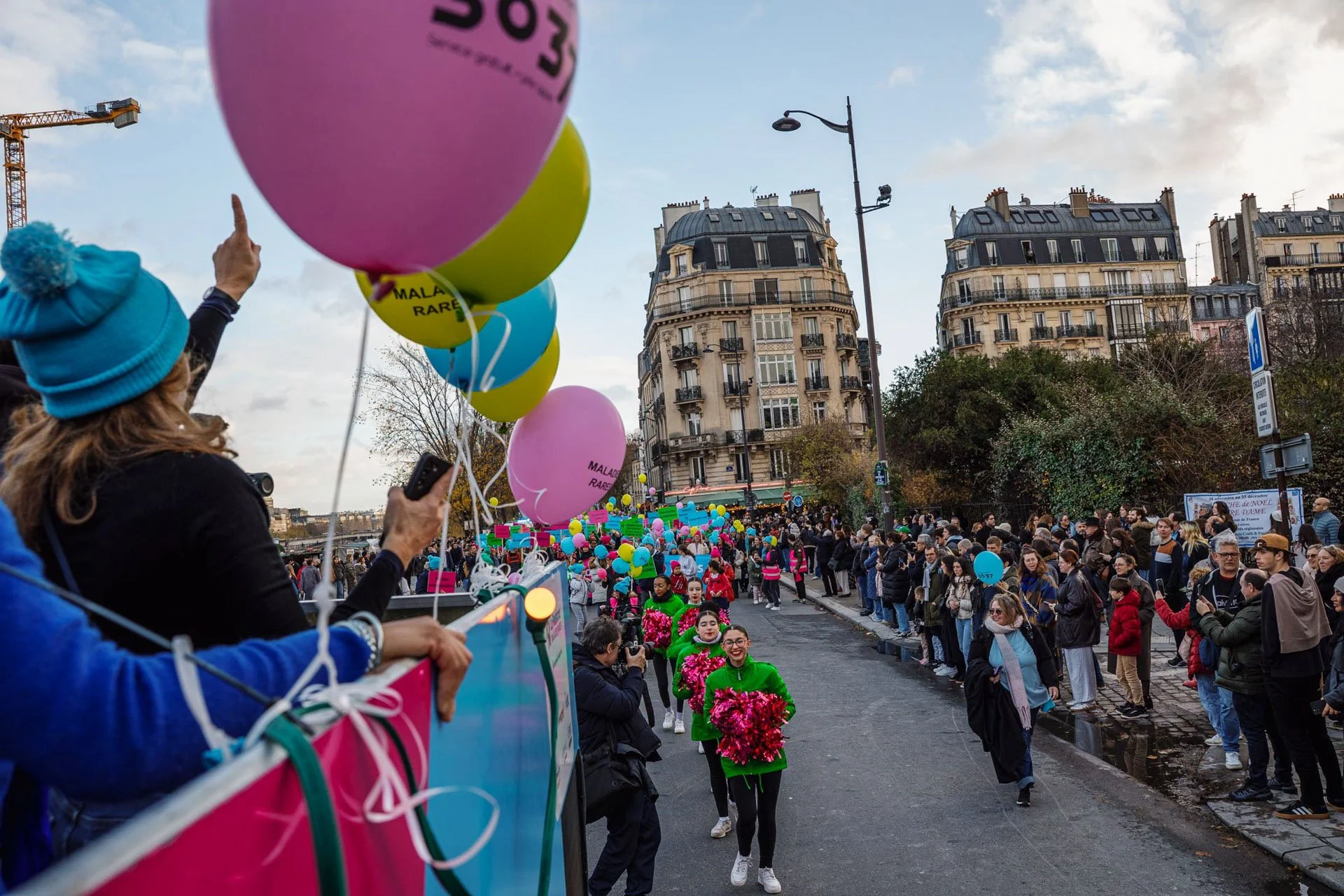 Une grande foule de personnes participent à un événement en plein air avec des ballons colorés, des pancartes, des crieurs d'encouragement et une vue sur des bâtiments parisiens en arrière-plan, probablement une marche ou une rassemblement pour une c