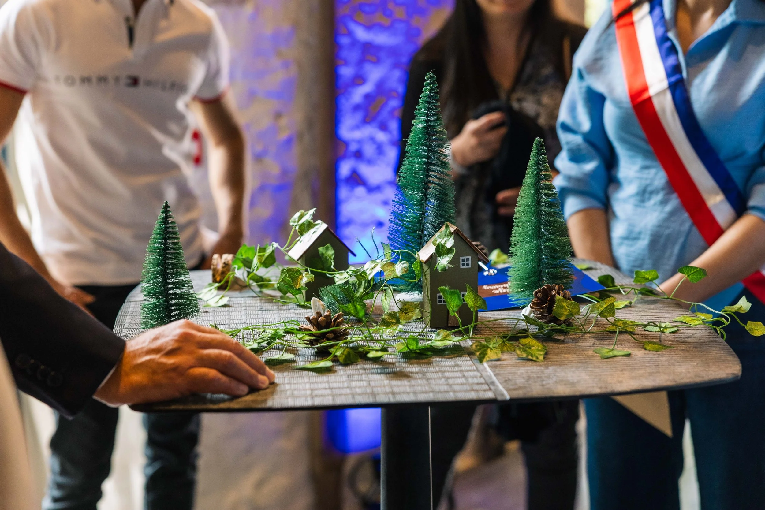 Groupe de personnes observant un projet de maquette de Noël avec des sapins en fil de fer, des petits modèles de maisons, des branches de feuilles vertes, des pommes de pin, sur une table.