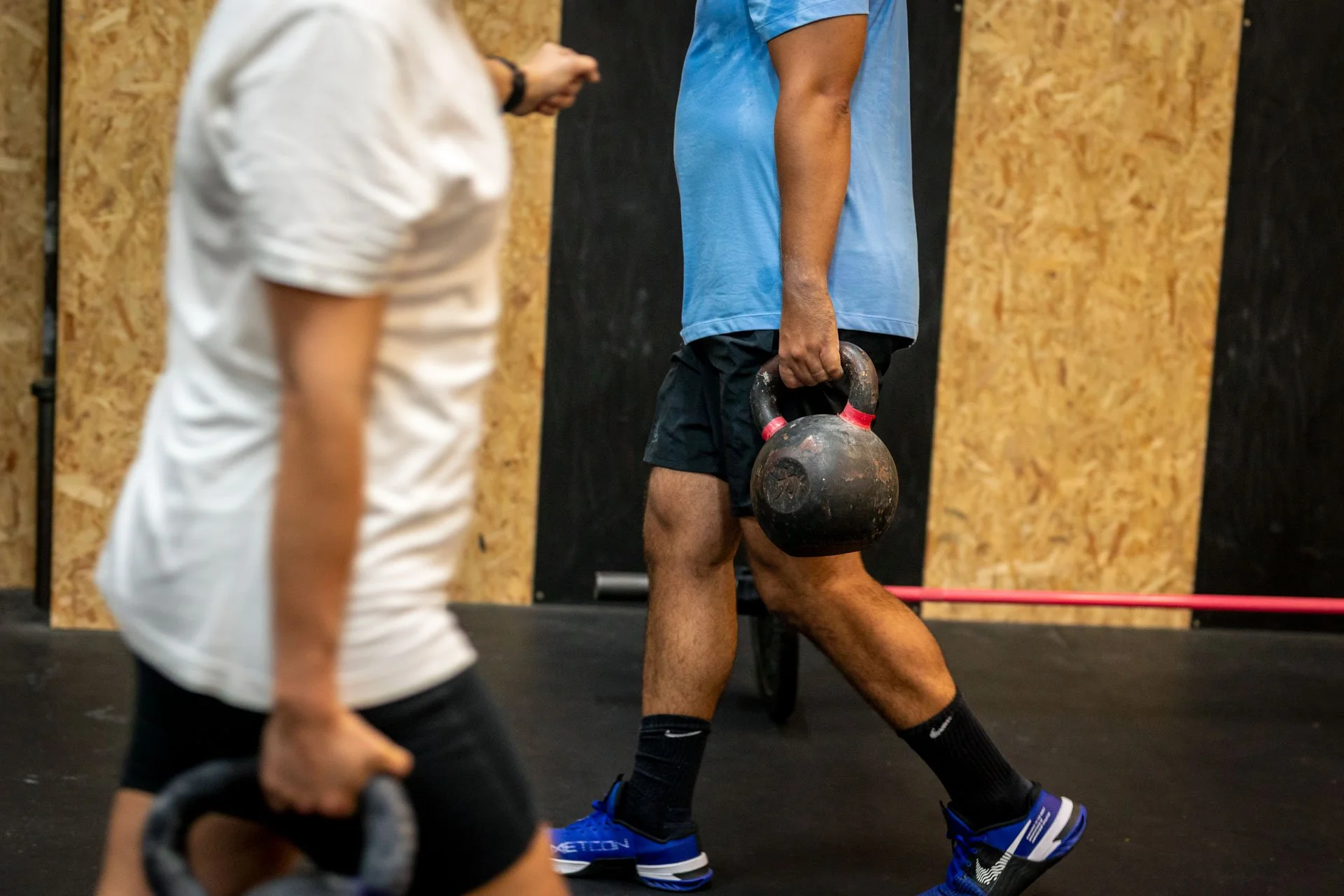 Un homme tenant un kettlebell en position de marche lors d'une séance d'entraînement en salle de sport. Personne en mouvement en premier plan. Mur en bois en arrière-plan.