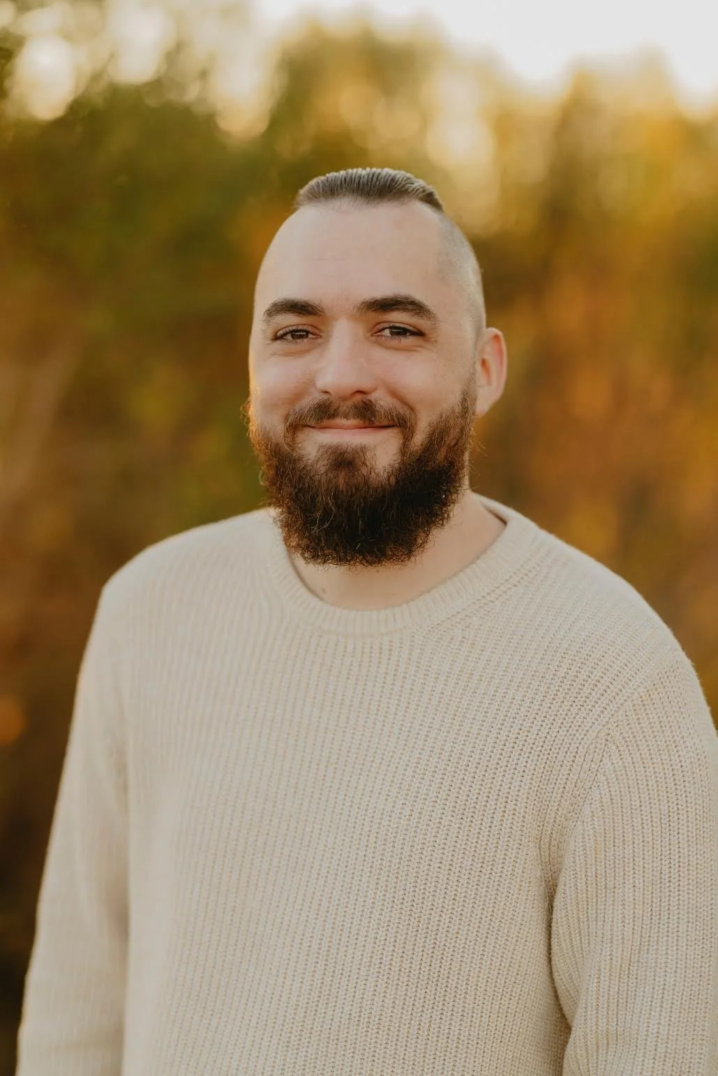A young man with a beard and mustache smiling outdoors during sunset. He is wearing a cream-colored sweater and has a short haircut, with a background of autumn-colored trees.