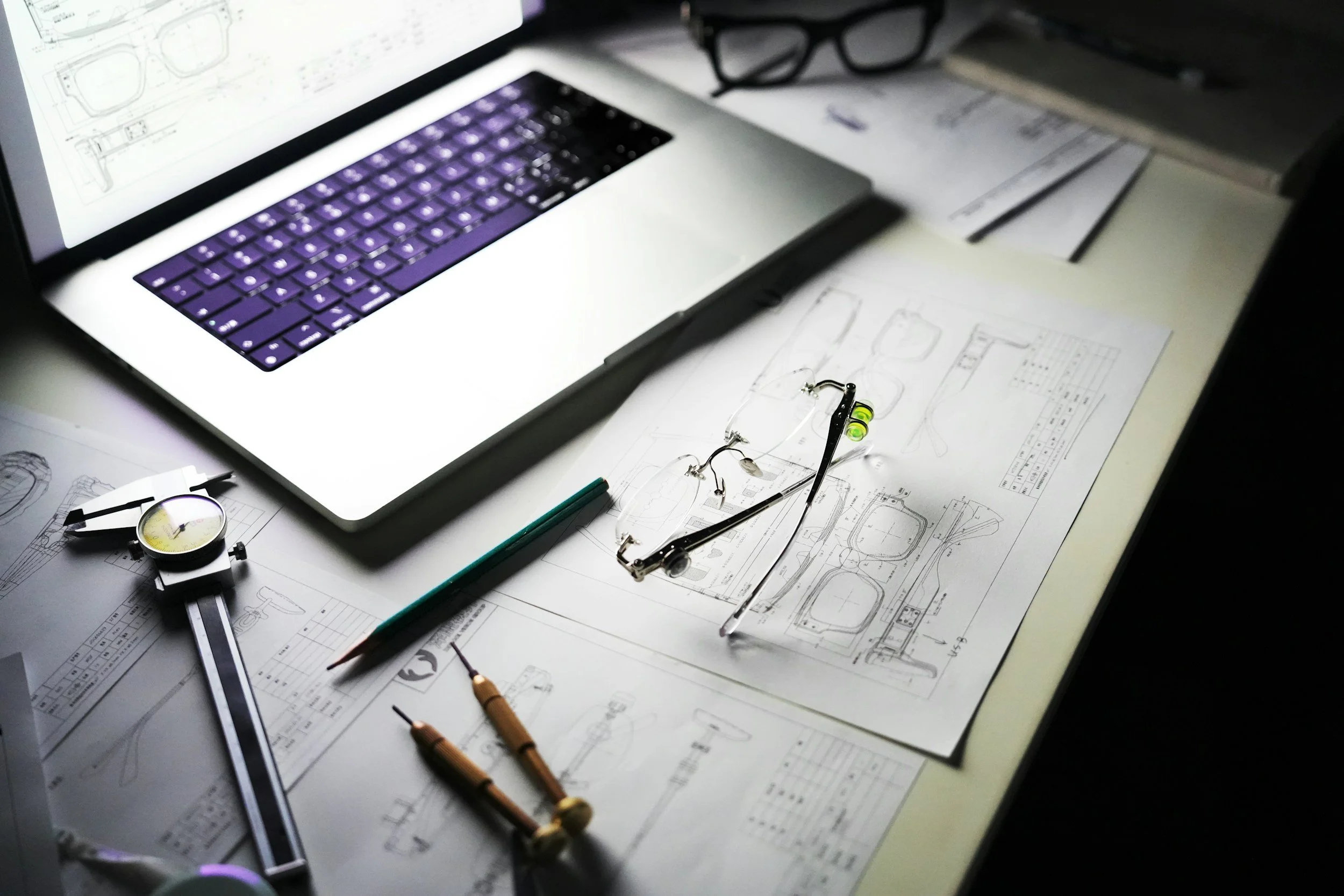 Architectural blueprints and technical drawings on a table with a pair of glasses, a compass, a ruler, and a pen, illuminated by a laptop screen displaying a digital design.