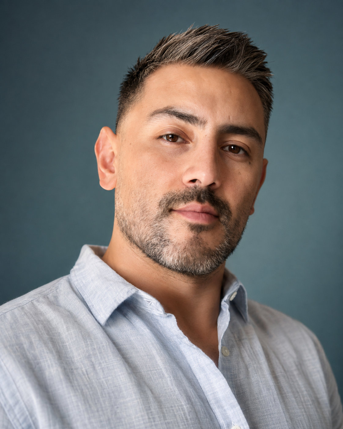 A close-up portrait of a man with short dark hair, a beard, and wearing a light gray button-up shirt, against a dark teal background.