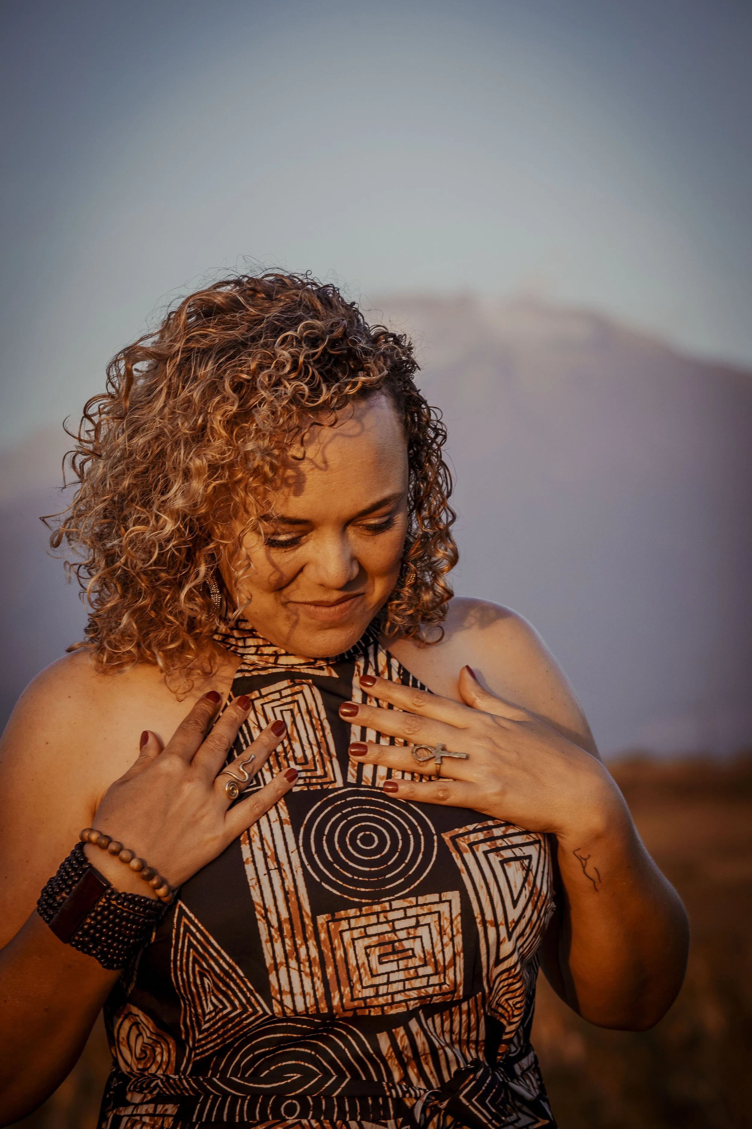 A woman with curly hair smiling and holding her hands over her chest in a patterned dress outdoors during sunset.