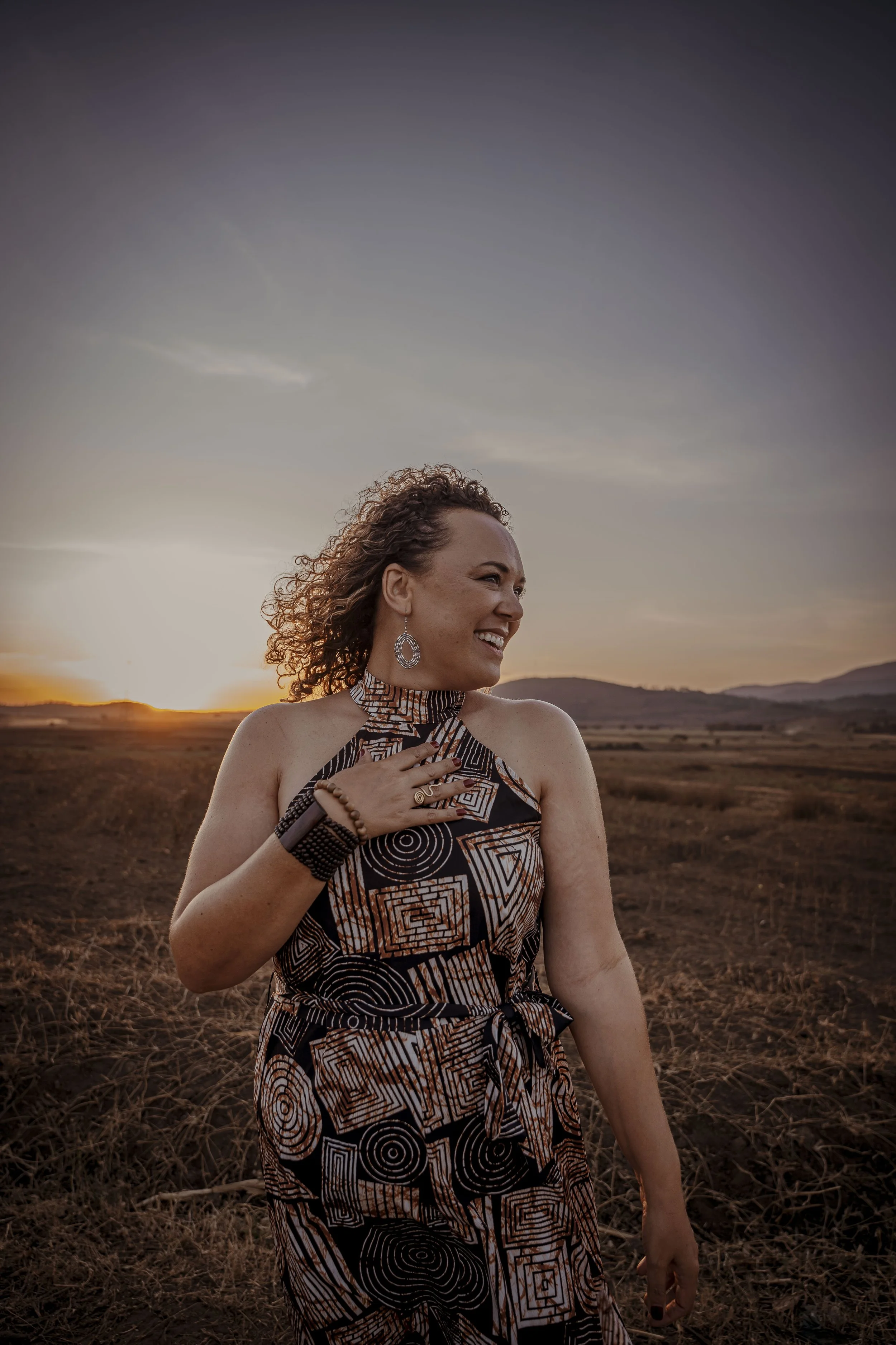A woman with curly hair wearing earrings and a patterned dress, standing in a field at sunset, with mountains in the background.