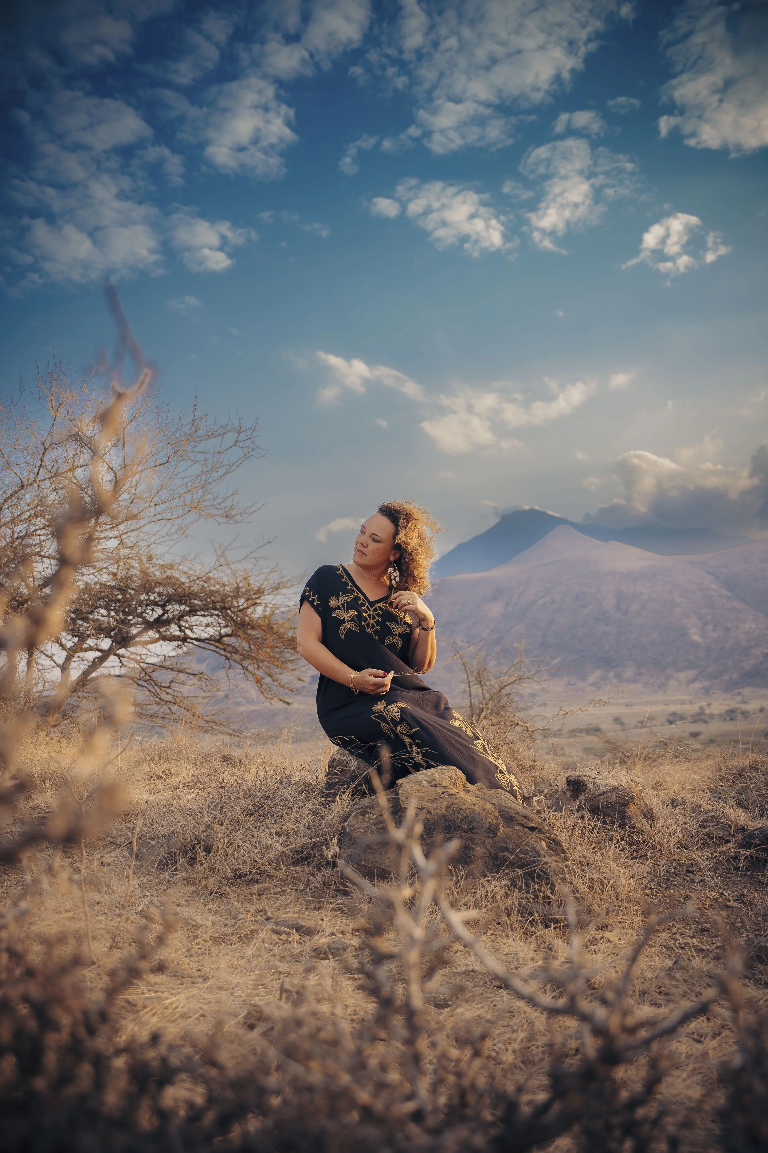 A woman with curly hair wearing a traditional black dress with gold embroidery, sitting on a rock in a dry, grassy field with a mountain and cloudy sky in the background.