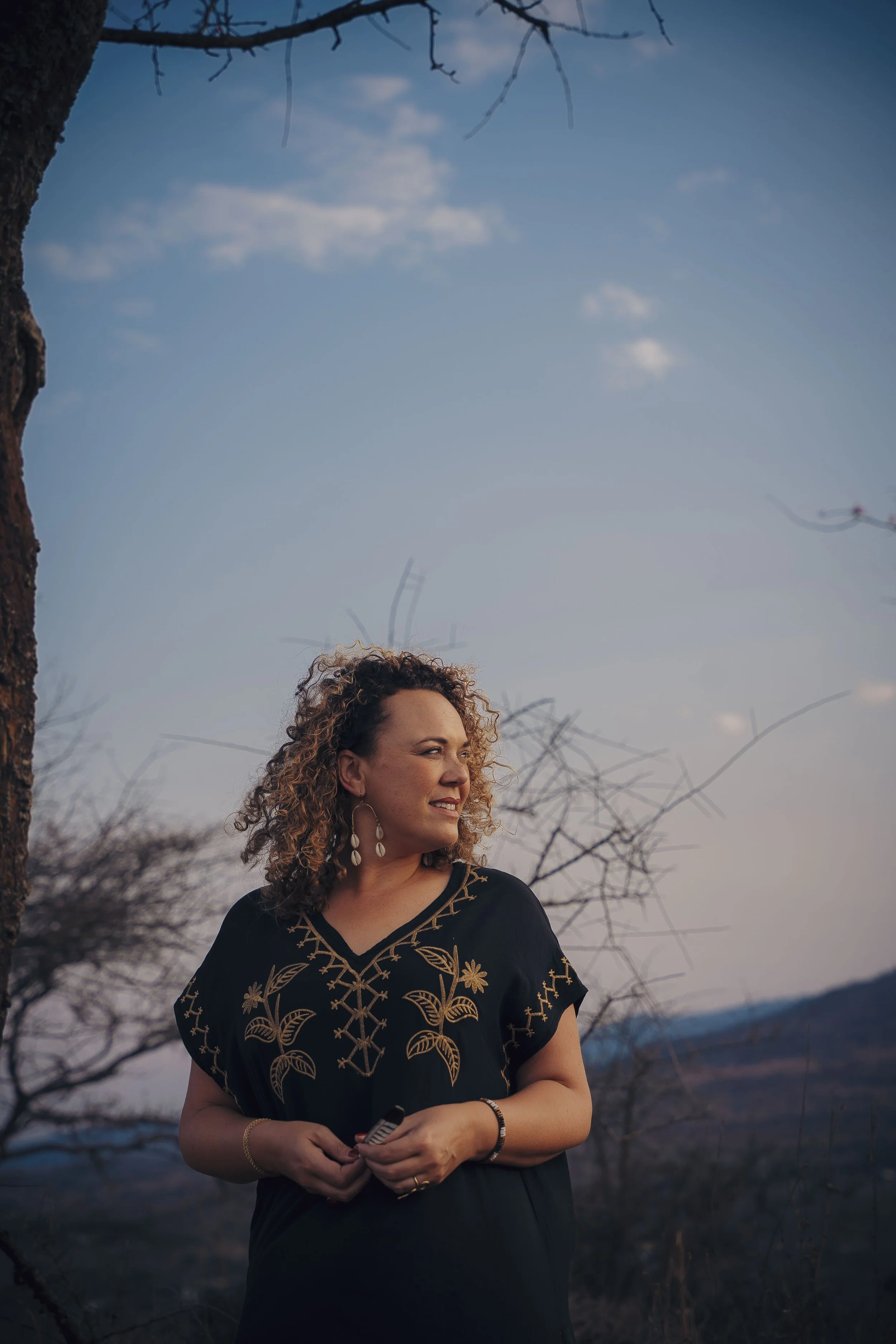 A woman with curly hair, wearing a black dress with floral gold embroidery and jewelry, stands outdoors near trees under a blue sky with clouds.