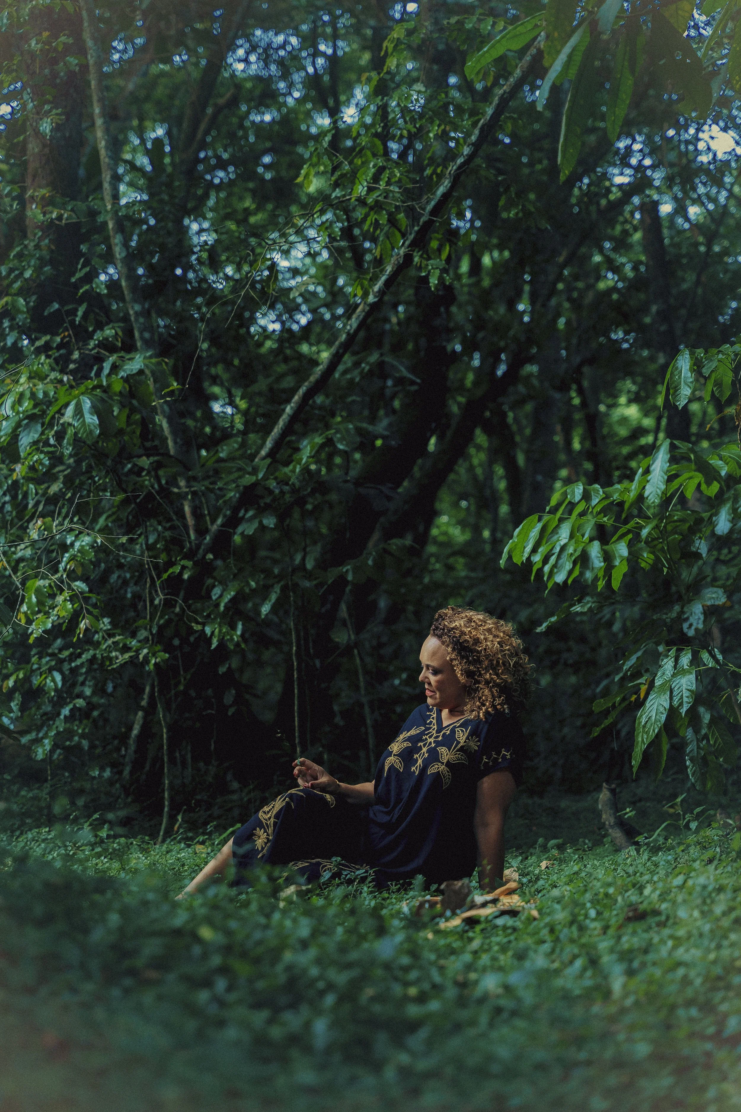 A woman with curly hair sitting in a forested area surrounded by lush green foliage.