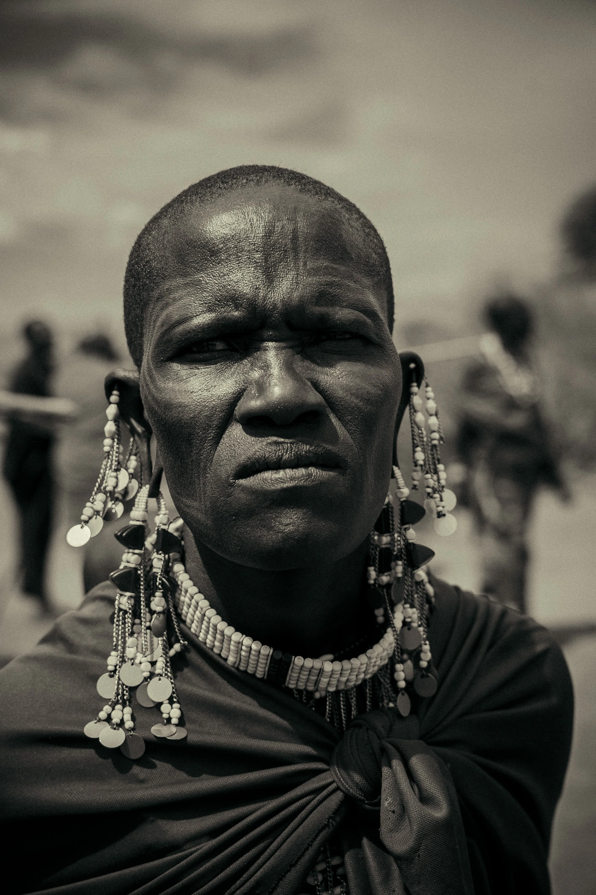 Close-up of an older African woman wearing traditional jewelry and clothing, with blurred background of people.