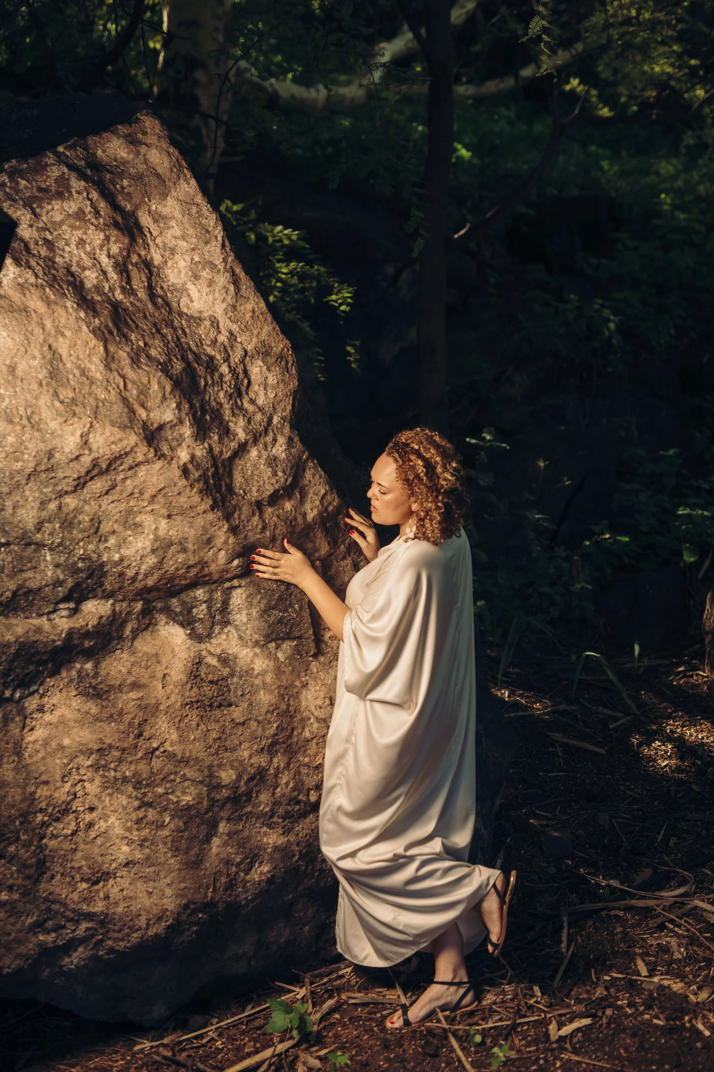 A woman with curly hair kneels in a forest, embracing a large boulder while wearing a flowing cream-colored dress and black high-heeled sandals.