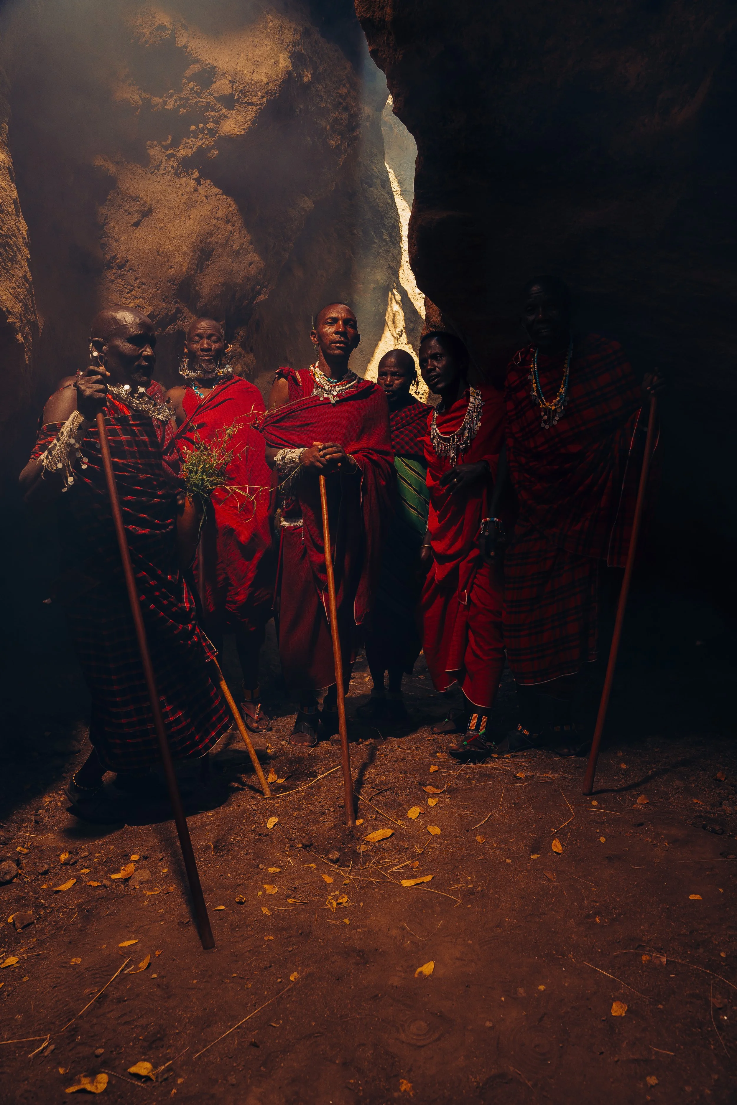 Group of Maasai tribesmen wearing traditional red shuka clothing and beaded jewelry, standing inside a cave with sunlight shining in from above, surrounded by brown rock formations.