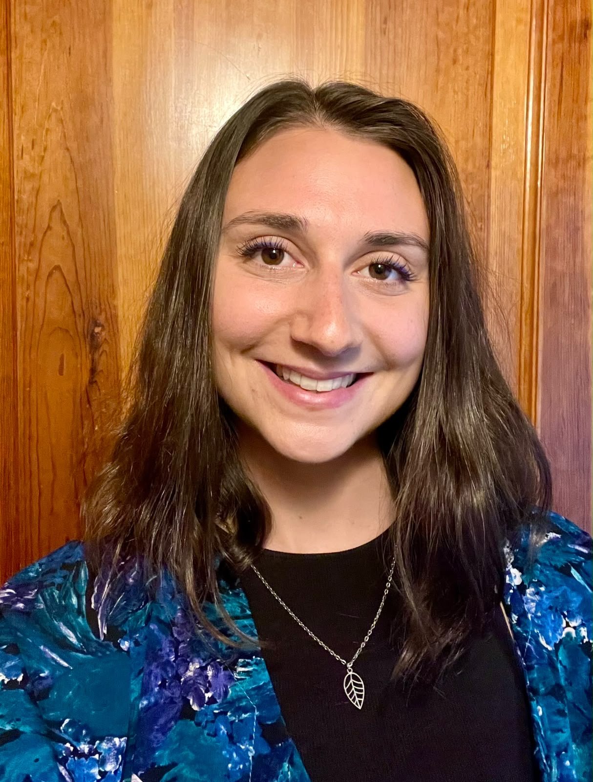 selfie of Maya Harvey smiling with teeth showing wearing colourful top and silver leaf necklace. Neutral background of brown wooden doorframe