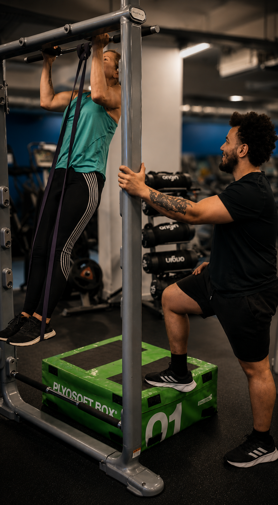 A woman doing pull-ups on a fitness rack with the assistance of a trainer at a gym. The trainer is holding onto the rack, providing support, while the woman is lifting herself up.