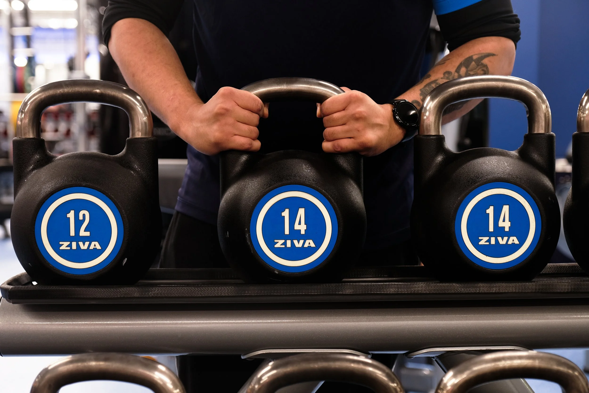 A person holding a kettlebell in a gym, with other kettlebells labeled with different weights on a rack in front of them.