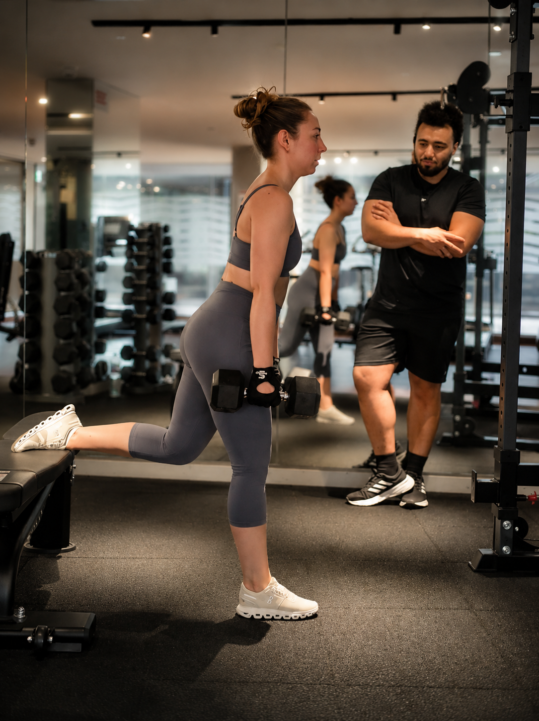 A woman in workout clothes performing a lunging exercise with a dumbbell while a trainer monitors her at a gym.