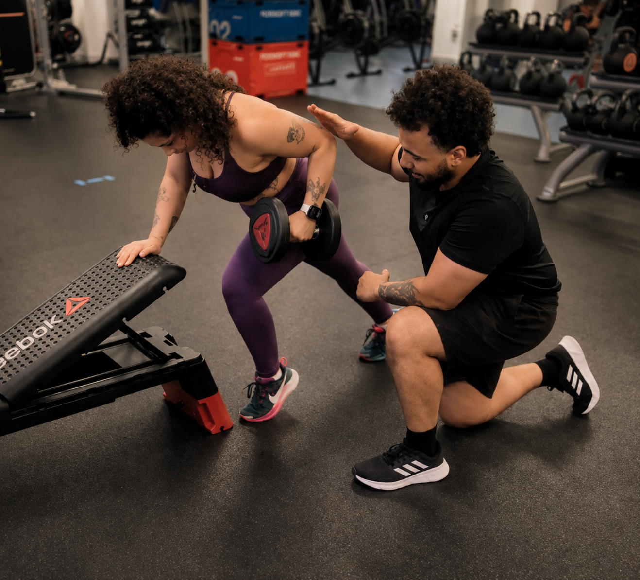 A woman performing a step-up exercise with dumbbells under the guidance of a trainer in a gym.