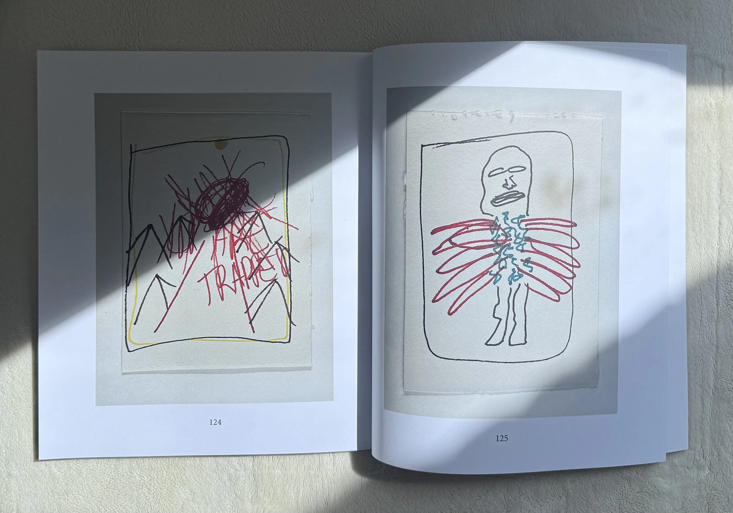 Open sketchbook with two pages displaying line drawings; the left page has a colorful, chaotic drawing with text reading 'TRAPPED', while the right page depicts a minimalistic figure with red wings and blue scribbles.