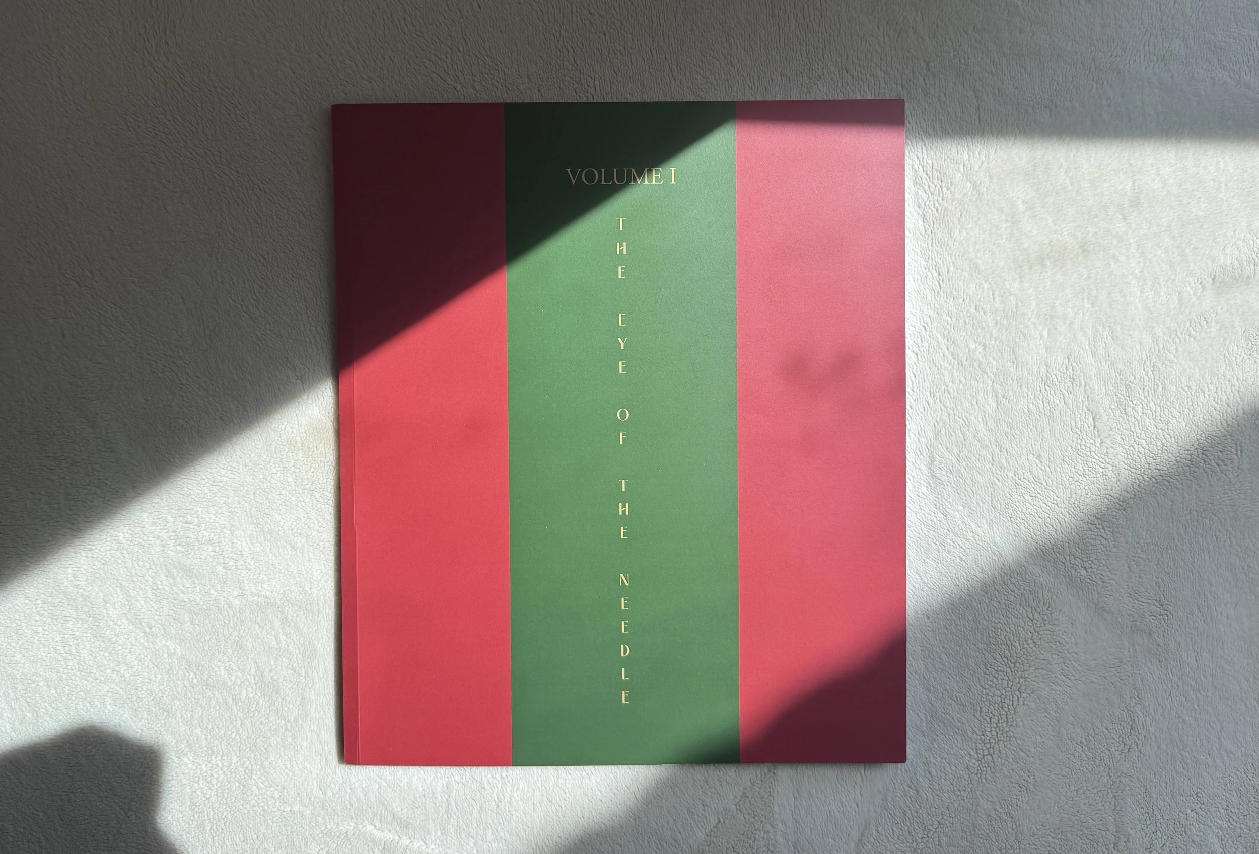 A book cover titled 'Volume I The Eye of the Needle' with a design featuring vertical red and green stripes and gold lettering, partially illuminated by sunlight on a white textured surface.