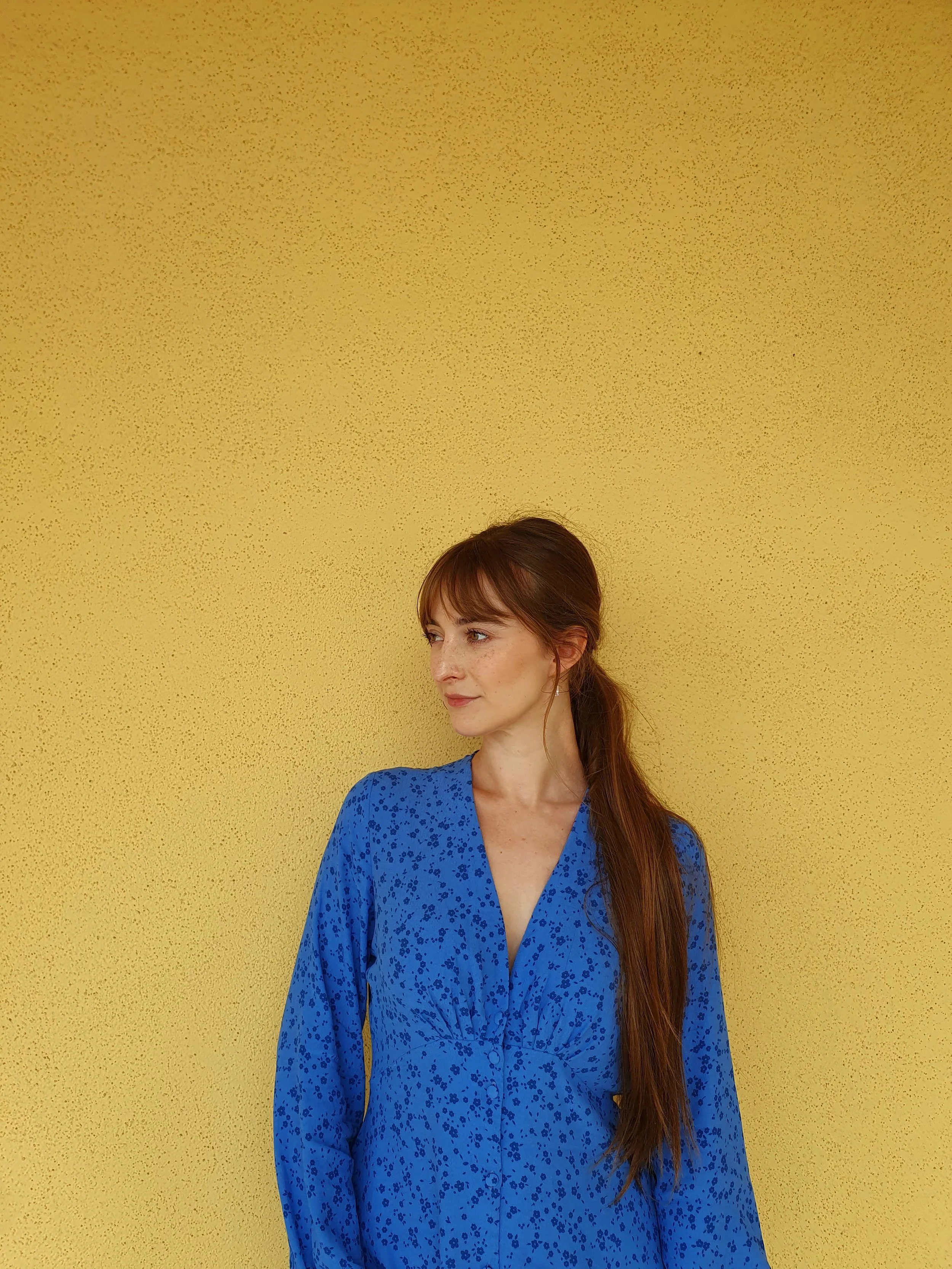 Woman with long red hair in a blue dress with small dark floral pattern standing against a yellow textured wall, looking to the side.
