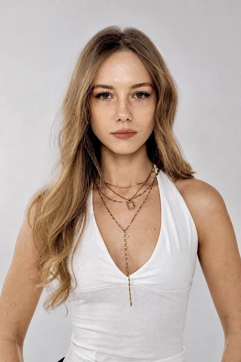 A woman with long, wavy, light brown hair wearing a white sleeveless top and layered gold necklaces looking directly at the camera with a neutral expression.