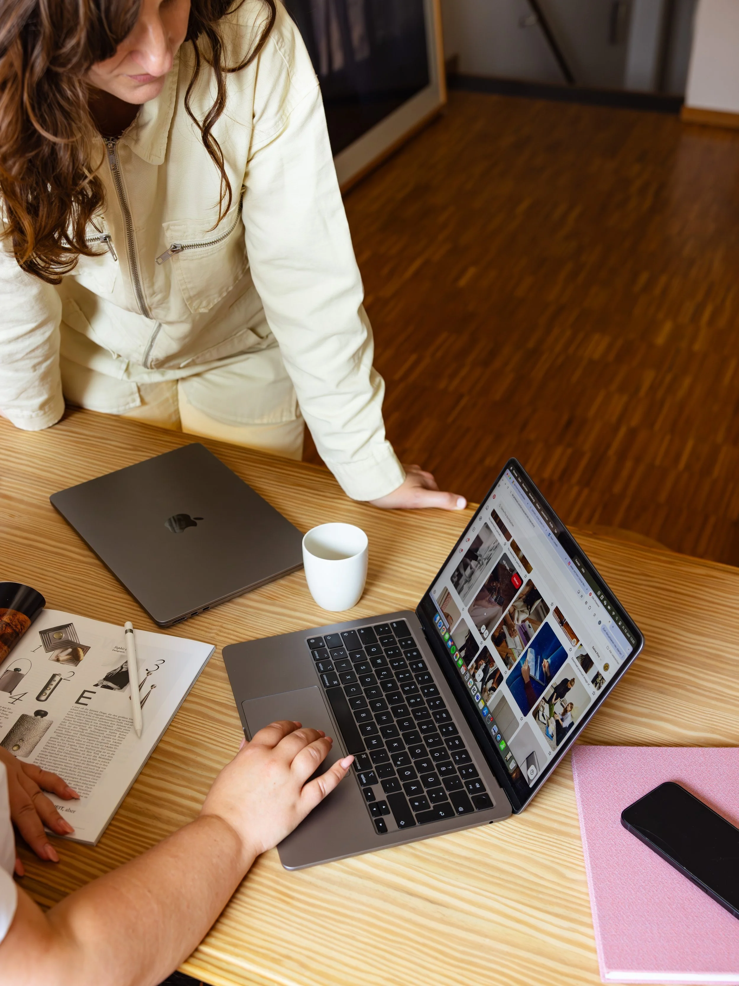 Zwei Personen arbeiten an einem Holzschreibtisch, einer benutzt einen Laptop, der andere schaut auf den Bildschirm, es gibt eine Tasse, ein Smartphone, ein Magazin und einen MacBook auf dem Tisch.