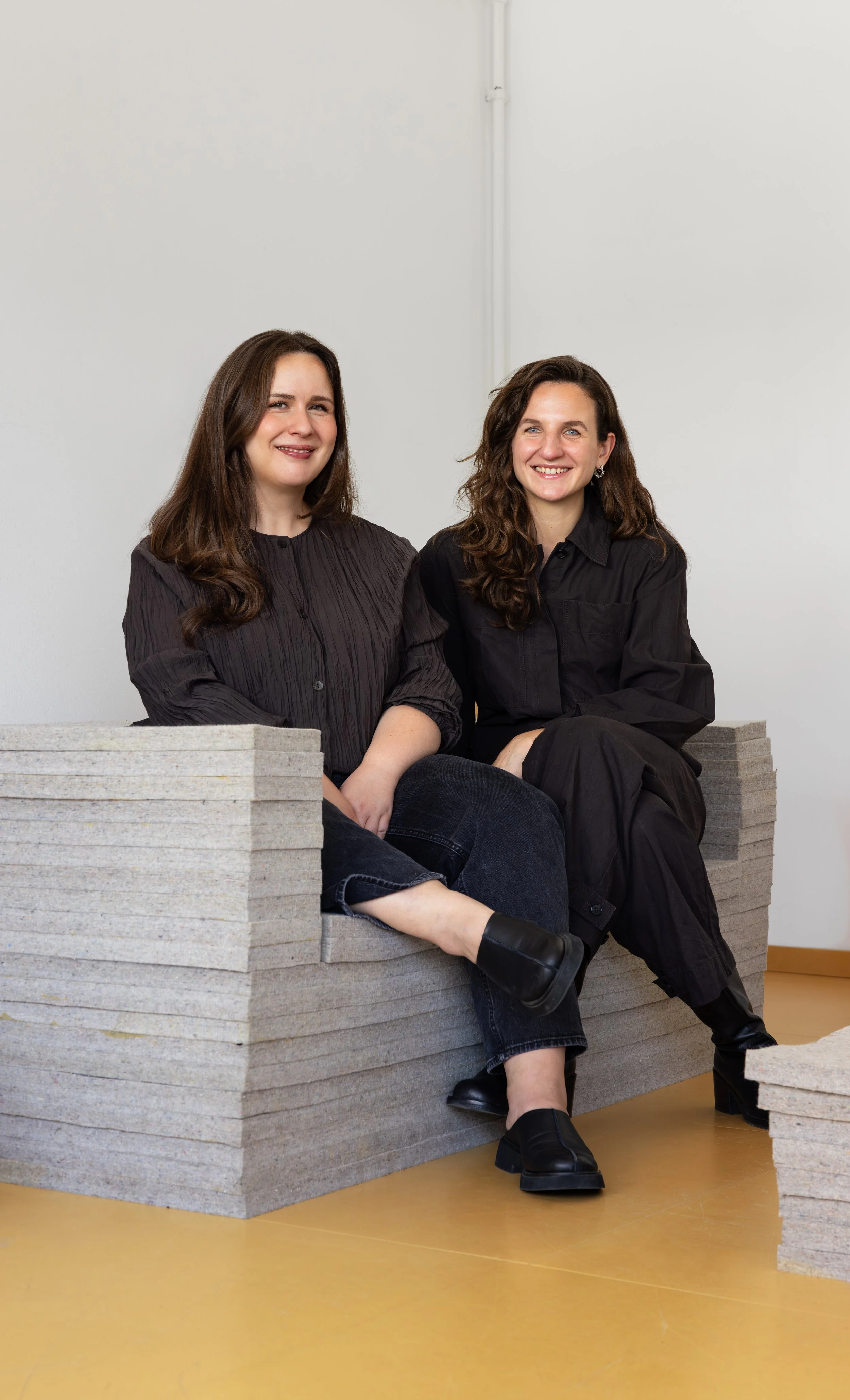 Zwei Frauen sitzen auf einer Sitzbank aus Pappe, lächeln und schauen in die Kamera, gegen einen weißen Hintergrund bei einem Foto- oder Medienstudio.