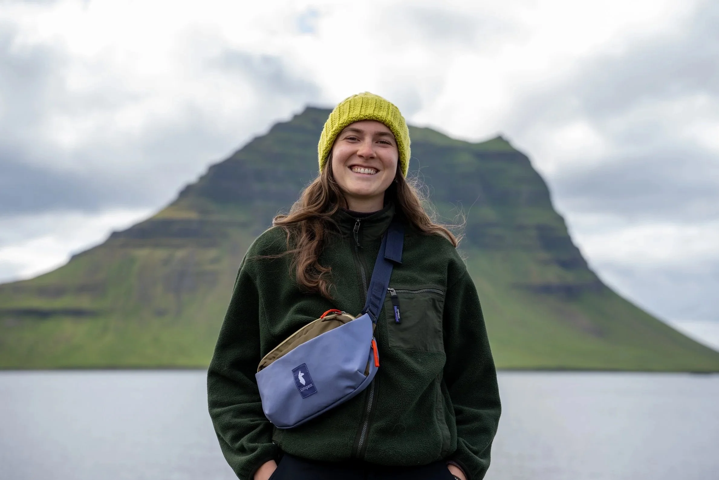 A woman with long brown hair wearing a yellow beanie and a black jacket, smiling at the camera, with a mountain in the background and cloudy sky.