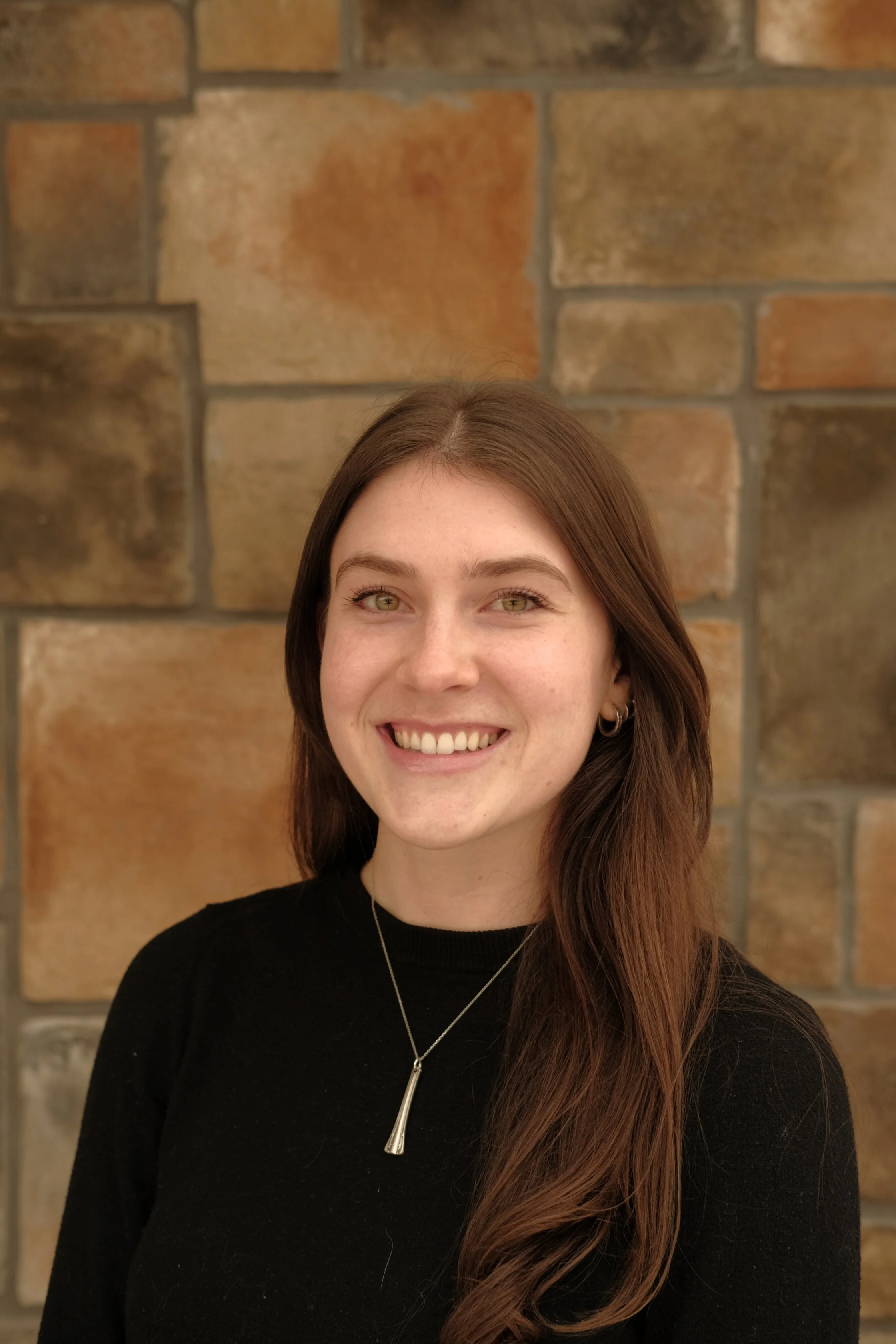 A young woman with long brown hair and a black top smiling, standing in front of a brick wall background.