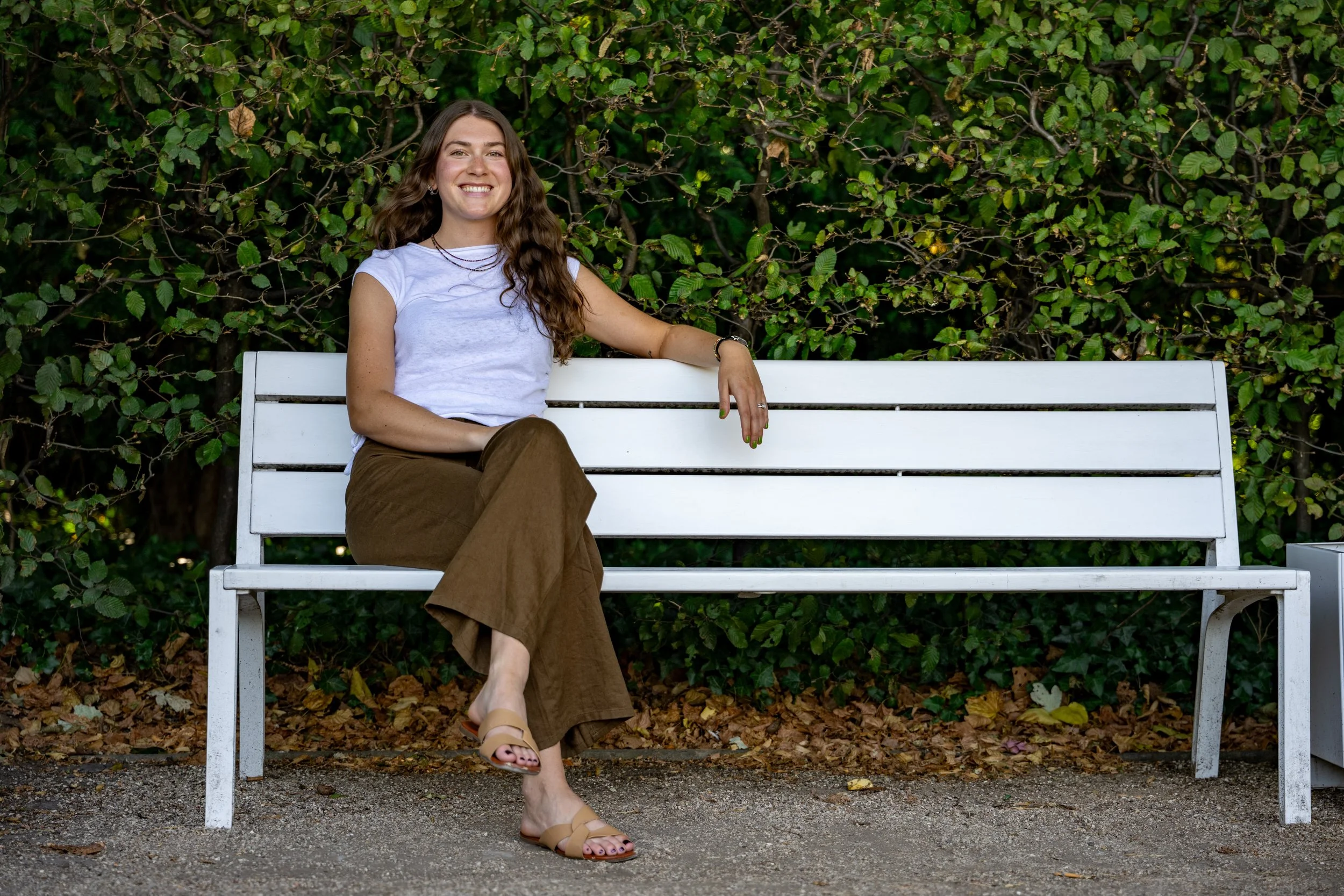 A young woman with long wavy brown hair sitting on a white park bench in front of a leafy green hedge, smiling at the camera, wearing a white sleeveless top, brown pants, and beige sandals.