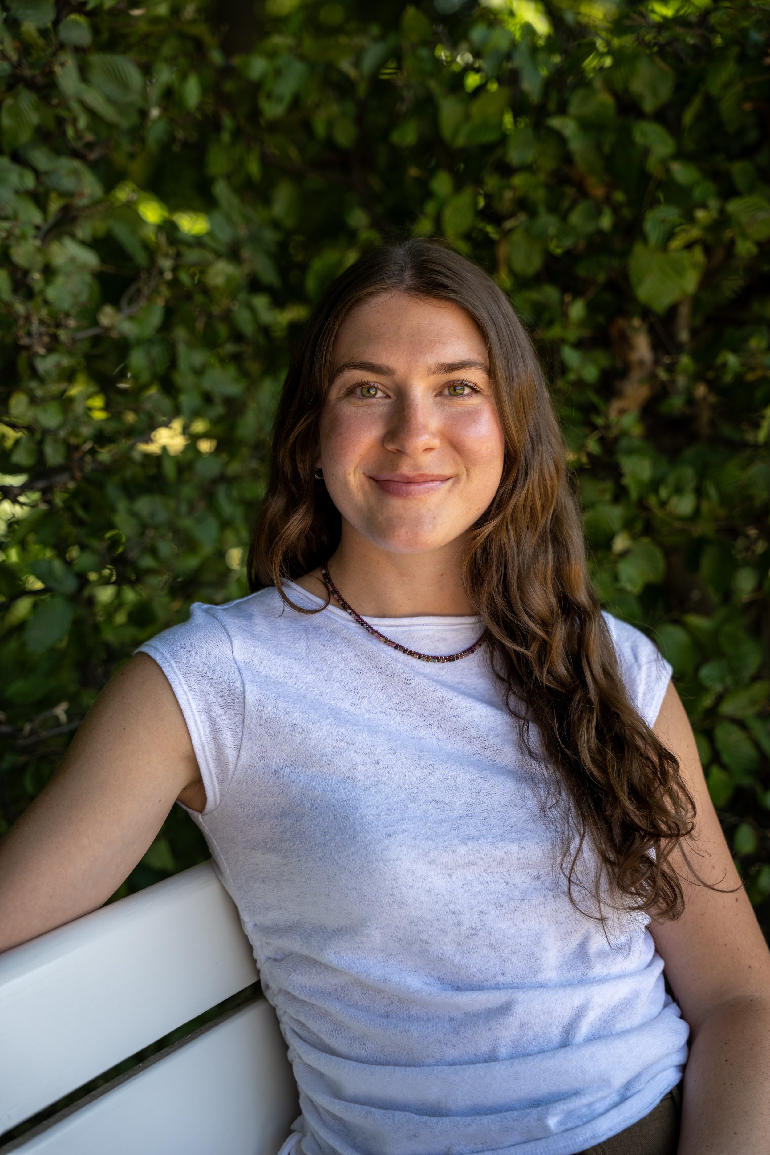 A young woman with long wavy brown hair and light skin, smiling, sitting on a white bench in front of green foliage, wearing a white T-shirt and a beaded necklace.
