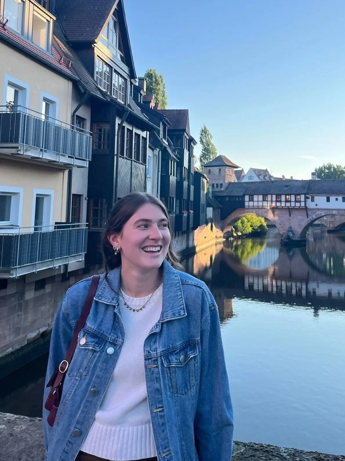 Smiling young woman with brown hair wearing a denim jacket and white top standing by a river with historic buildings and a stone bridge reflected in the water, under a clear blue sky.