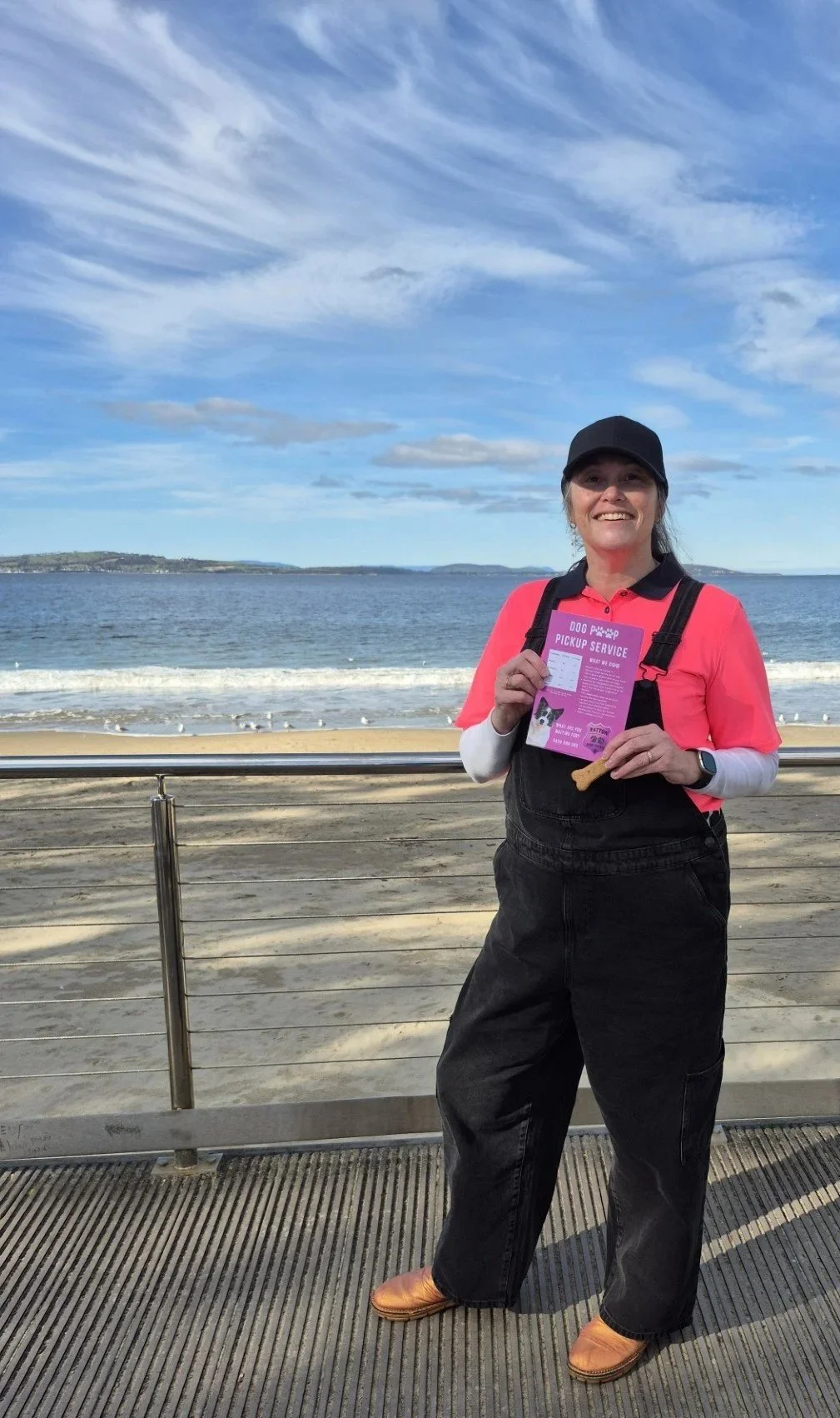 A woman smiling at the camera standing on a wooden pier with a beach and ocean in the background, holding a flyer and a dog biscuit, wearing a black cap, pink shirt, black overalls, and orange shoes.