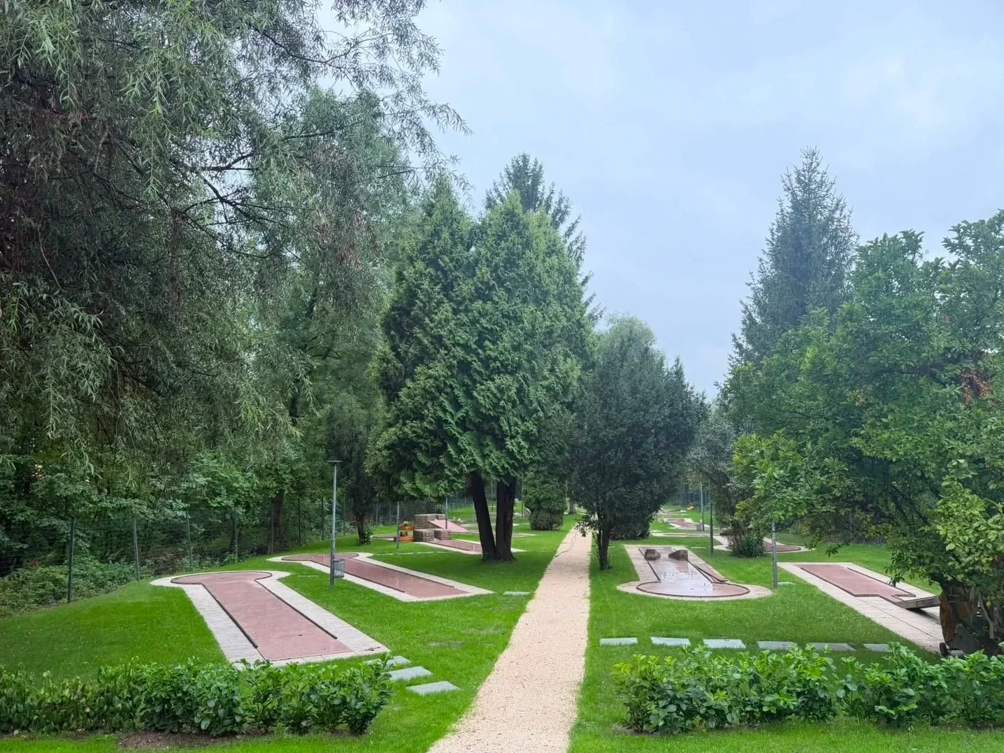 Ein gepflegter Park mit einer gepflasterten Wege, grünen Rasenflächen, Bäumen und mehreren Brunnen. Es gibt auch Laternen und Bänke.