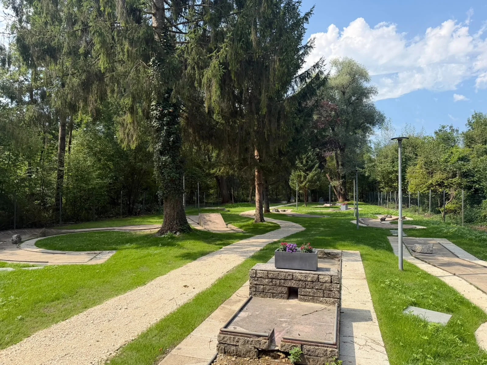 Einen gepflegten Park mit einem Weg, Bäumen, Pflanzen, Lampen und Minigolfbahnen bei gutem Wetter.