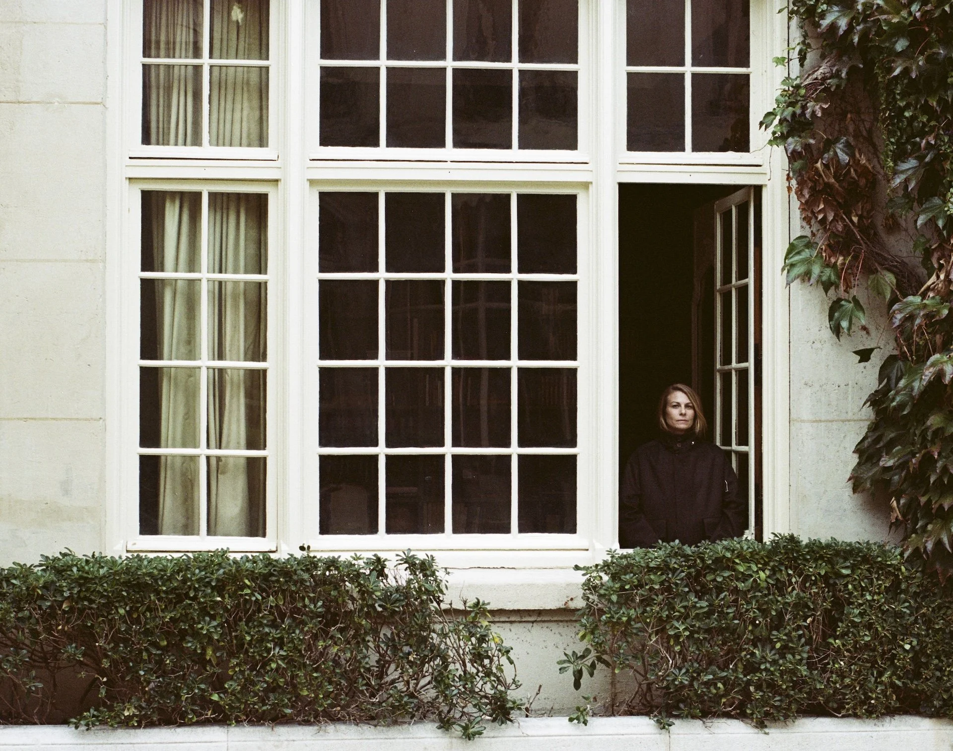 A woman with brown hair, wearing a dark jacket, standing in an open window with a grid of four over four panes. The window is part of a building with a beige stone exterior. There are green shrubs at the base of the window and some climbing ivy on the right side of the window.