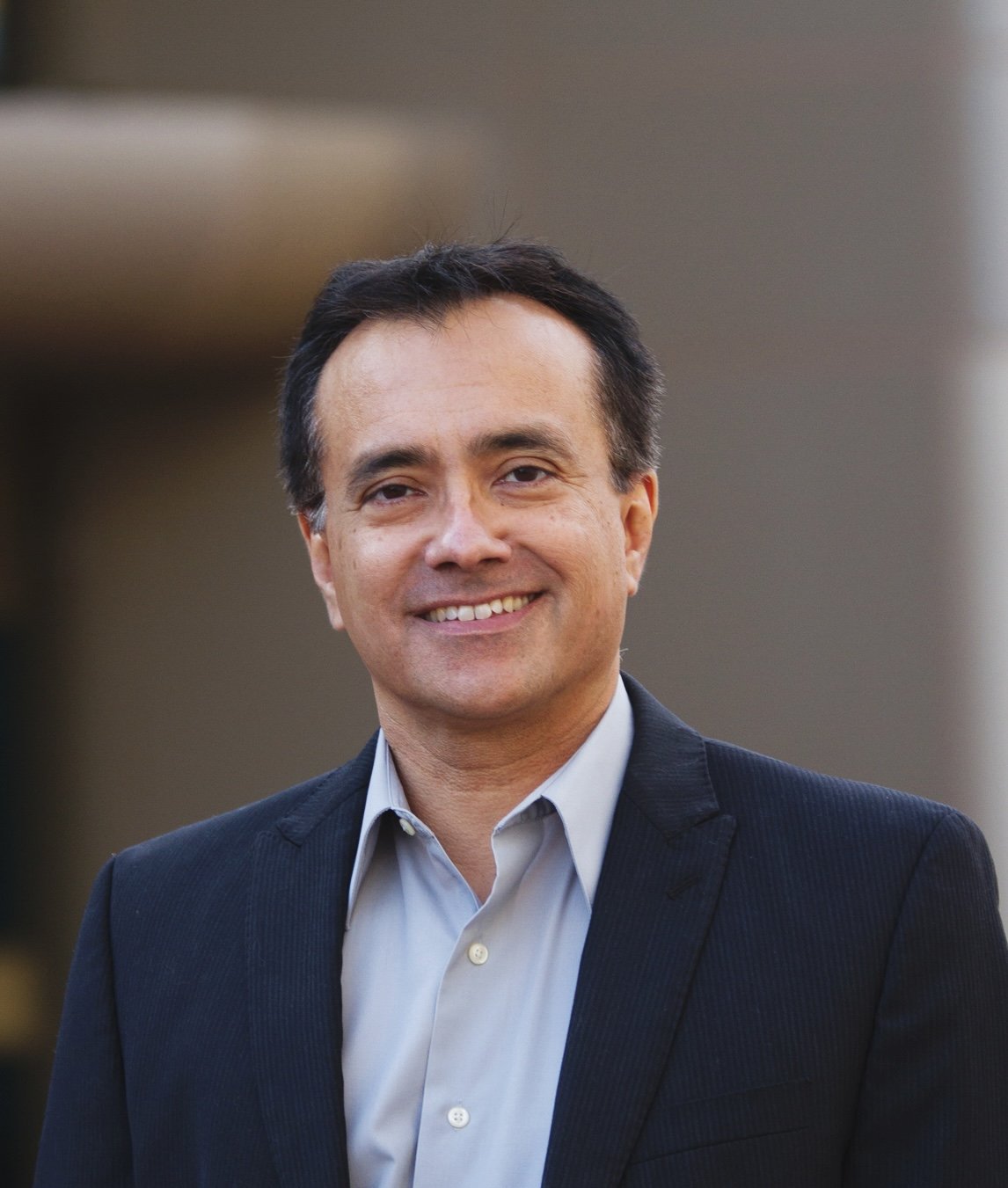 A man with dark hair and light skin, wearing a gray suit jacket and a white dress shirt, smiling in an outdoor setting.