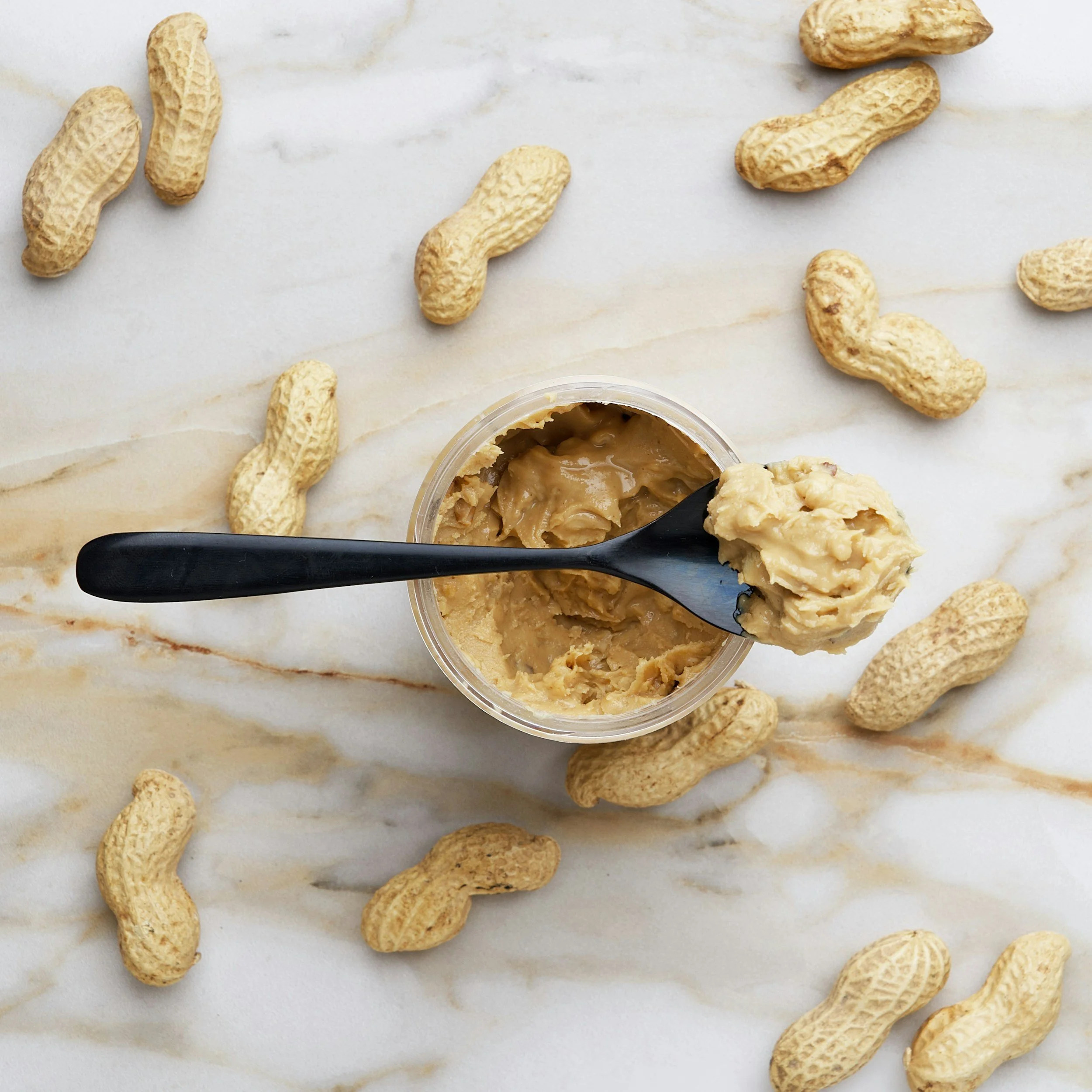 A clear plastic container of peanut butter surrounded by scattered unshelled peanuts on a white marble surface.