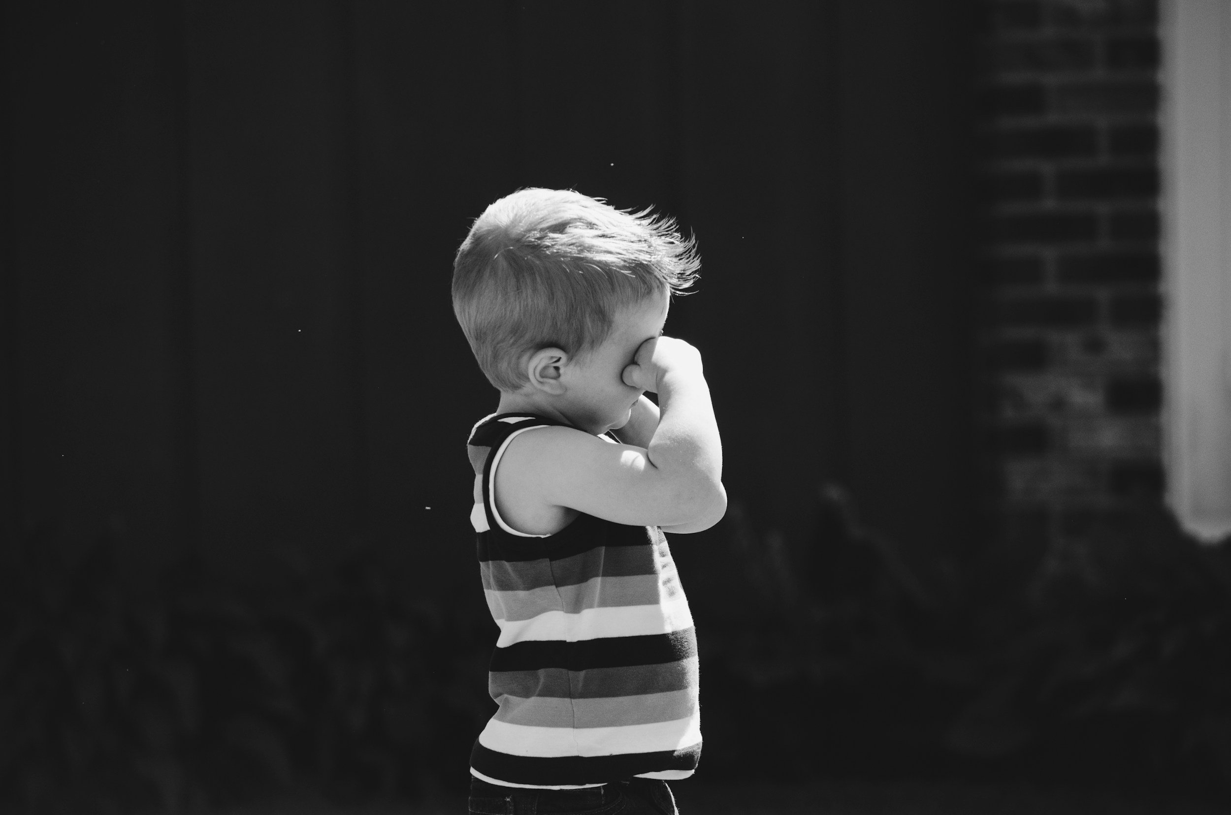 A young boy with short, light-colored hair, wearing a striped sleeveless shirt, stands outdoors with his eyes closed, covering his nose with his right hand. The background is dark with a large brick wall on the right.