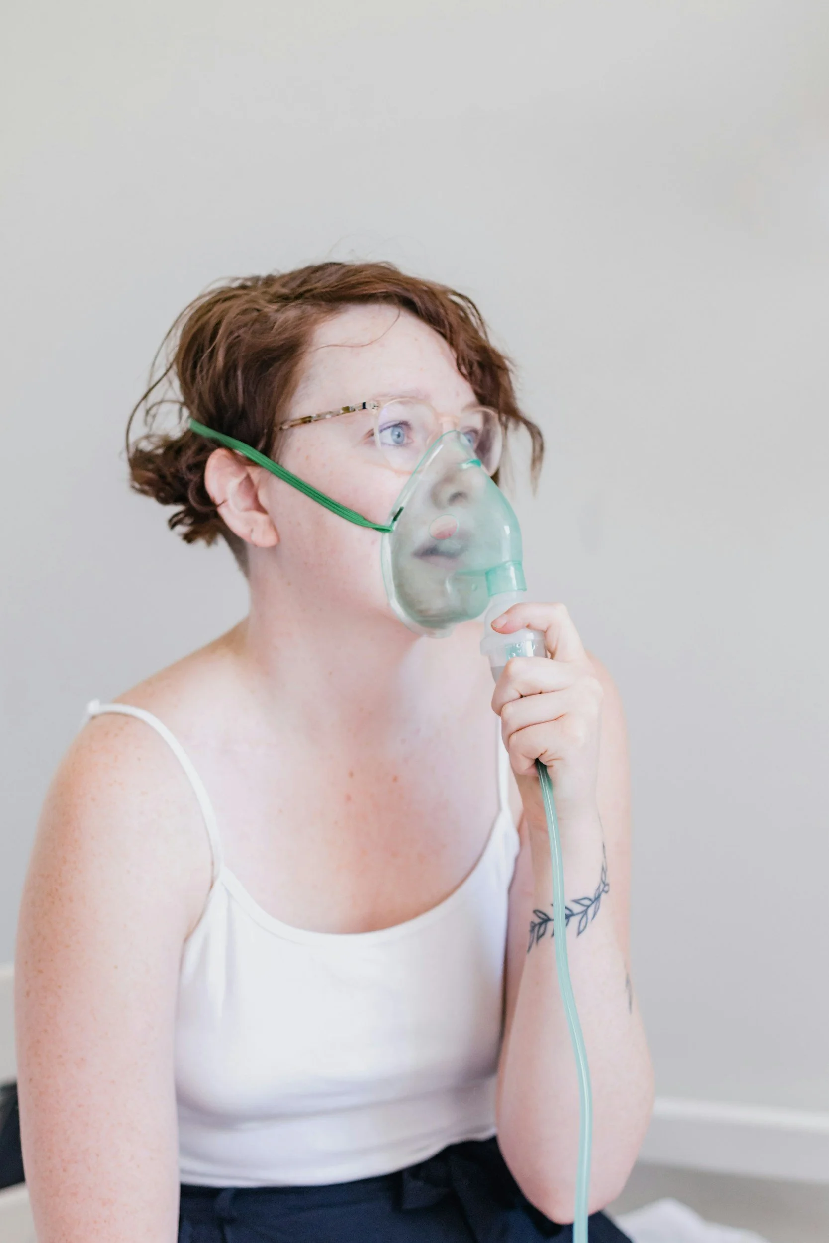 A woman with short to medium red hair, freckles, and blue eyes is holding a nebulizer mask to her face, wearing glasses, a white tank top, and has a tattoo on her left arm. The background is plain and light-colored.