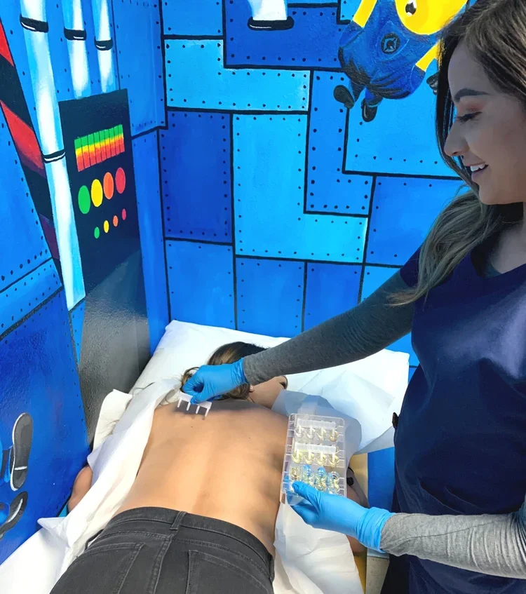 A healthcare professional in gloves preparing to give an injection to a patient lying face down on a bed in a medical setting with blue, rocket ship-themed walls.