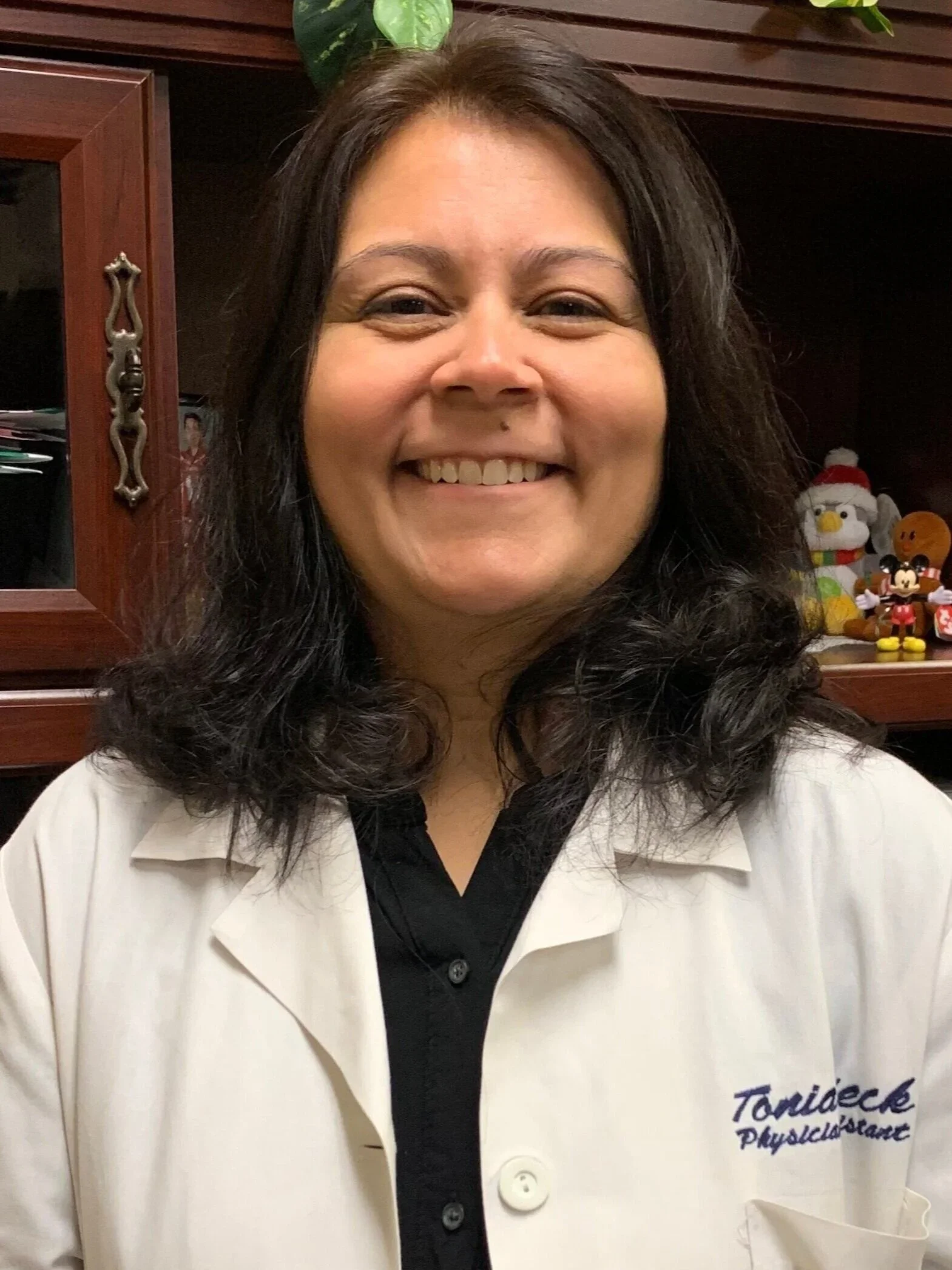 A woman with dark, curly hair smiling, wearing a white coat with a logo that reads 'Tomićke Physiotherapy.' In the background, there is a wooden cabinet with decorative handles and a couple of small plush toys.