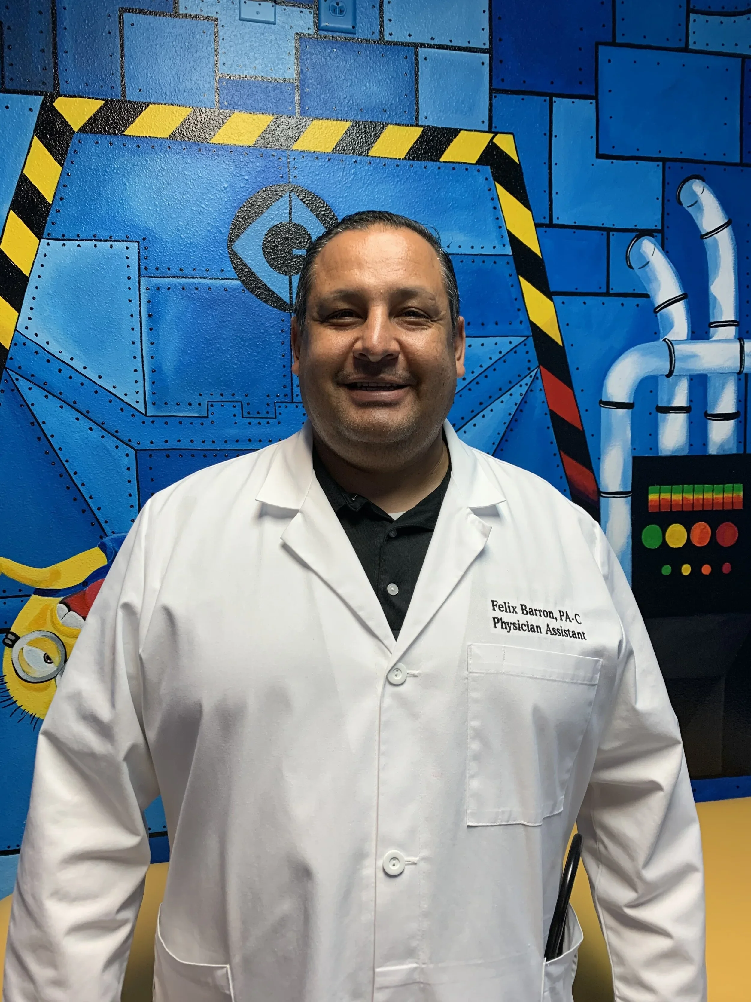 A man smiling, wearing a white lab coat with a name tag that reads 'Felix Barron, PA-C, Physician Assistant,' standing in front of a colorful mural with blue bricks, pipes, and a robot face.